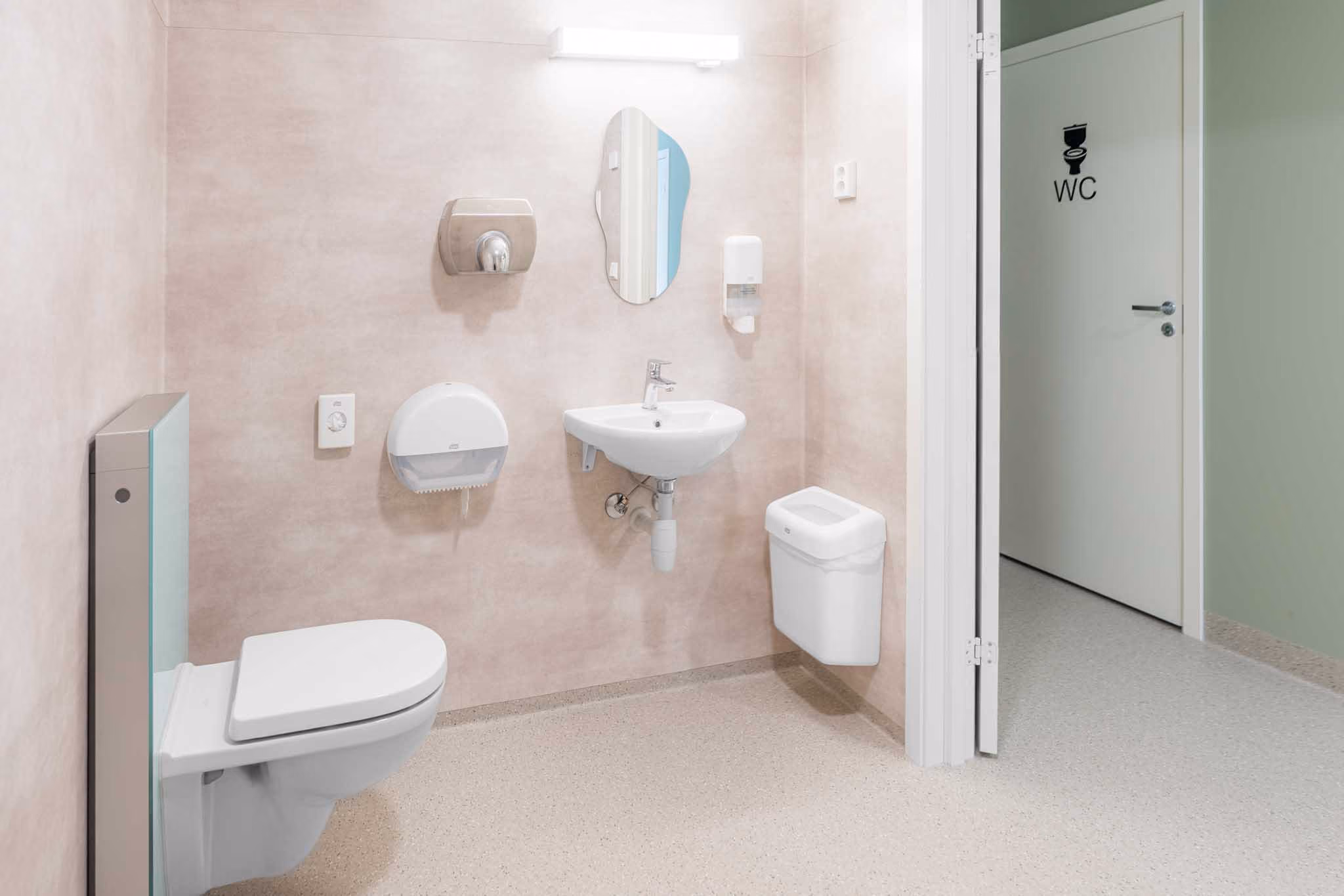 Rent toalett med veggmontert toalett, servant, speil og dispensere, sett mot WC-dør.