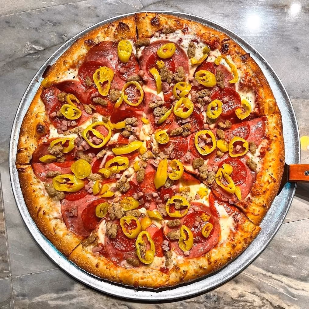 Whole pizza with pepperoni, sausage, ham, and banana pepper slices on a thin crust in a metal pan.