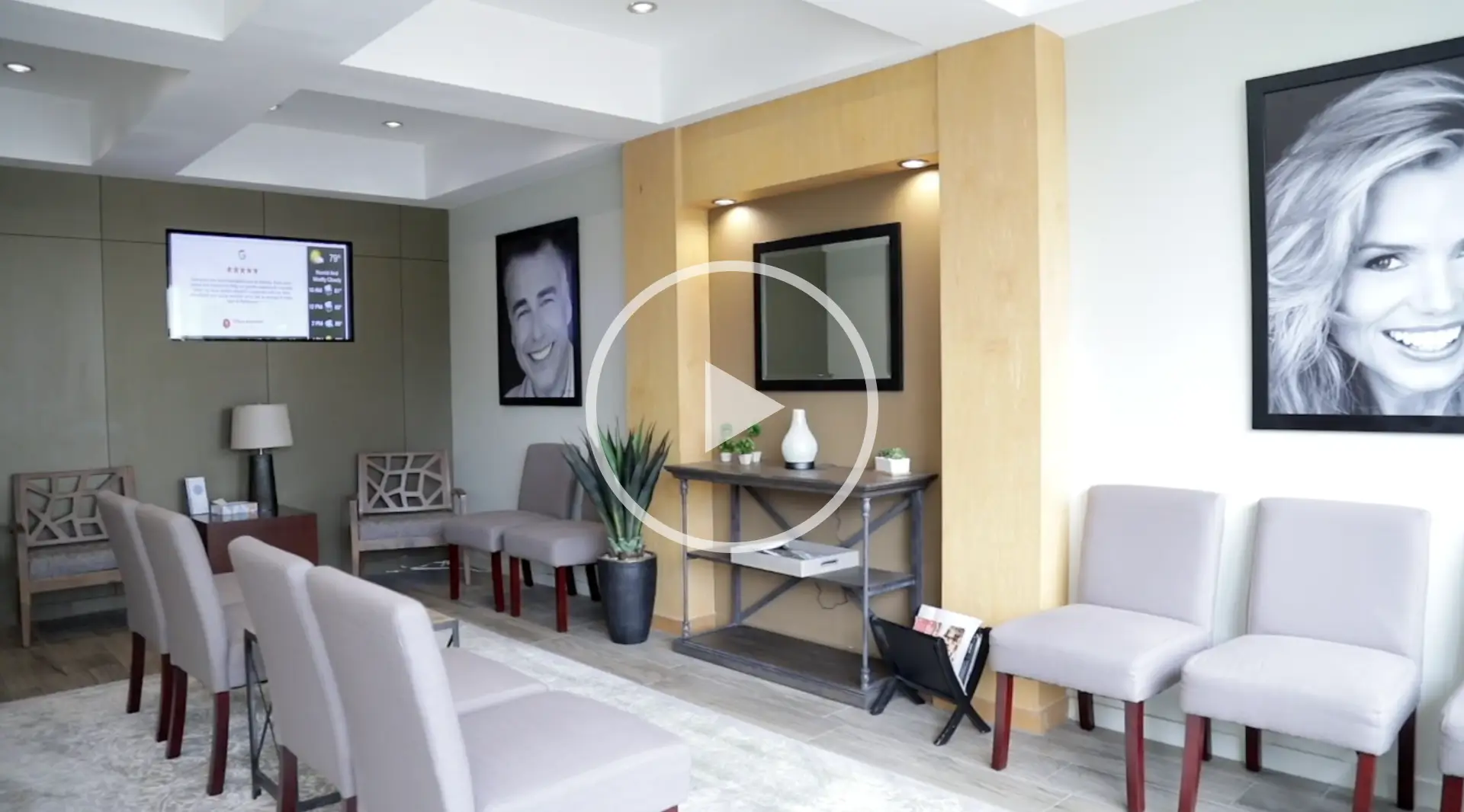 Waiting room with gray chairs, wall art, and plants. Room has a TV and light wood accents.