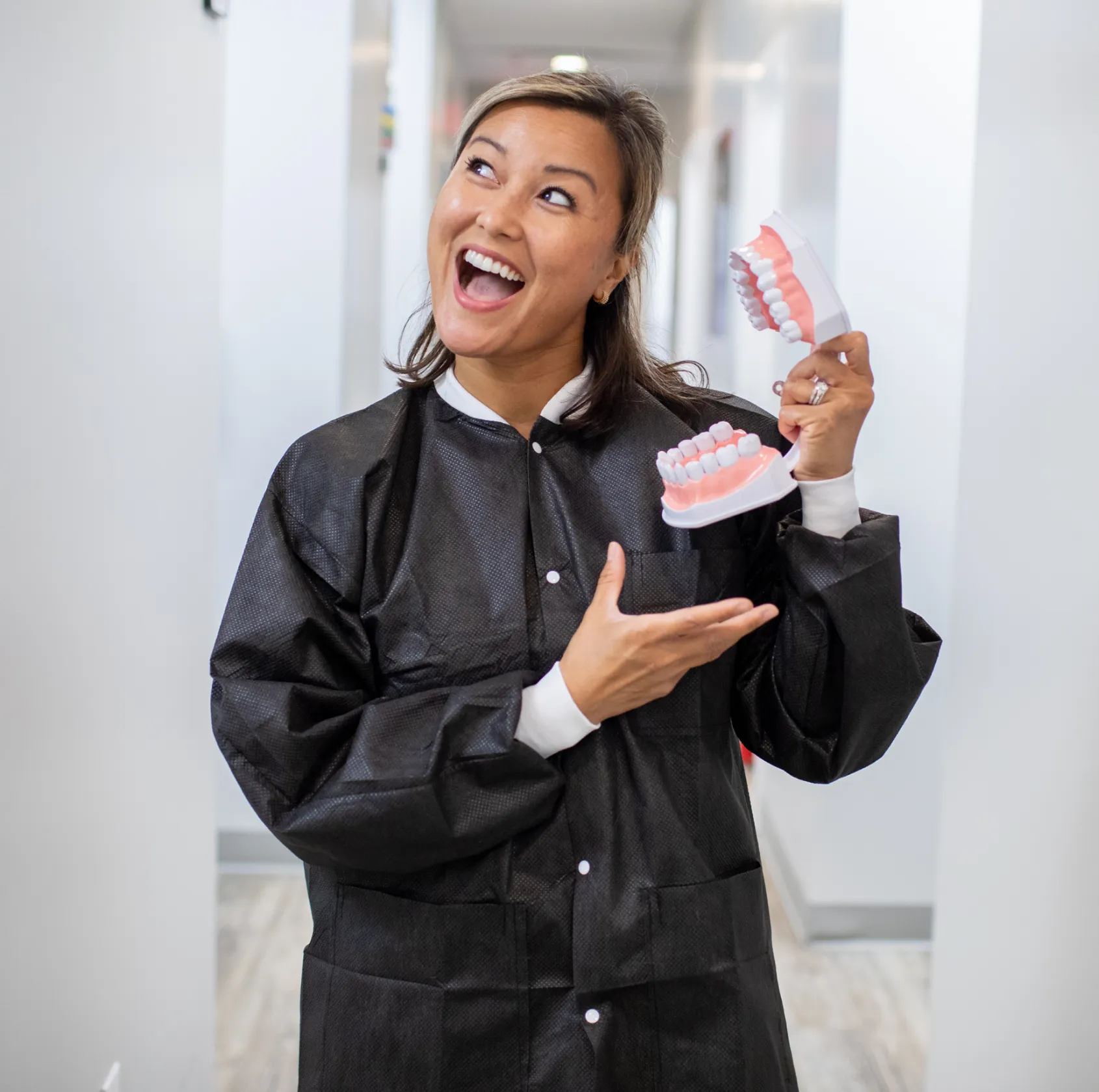 A woman in a dental office smiles and holds a model of teeth, pointing at it.