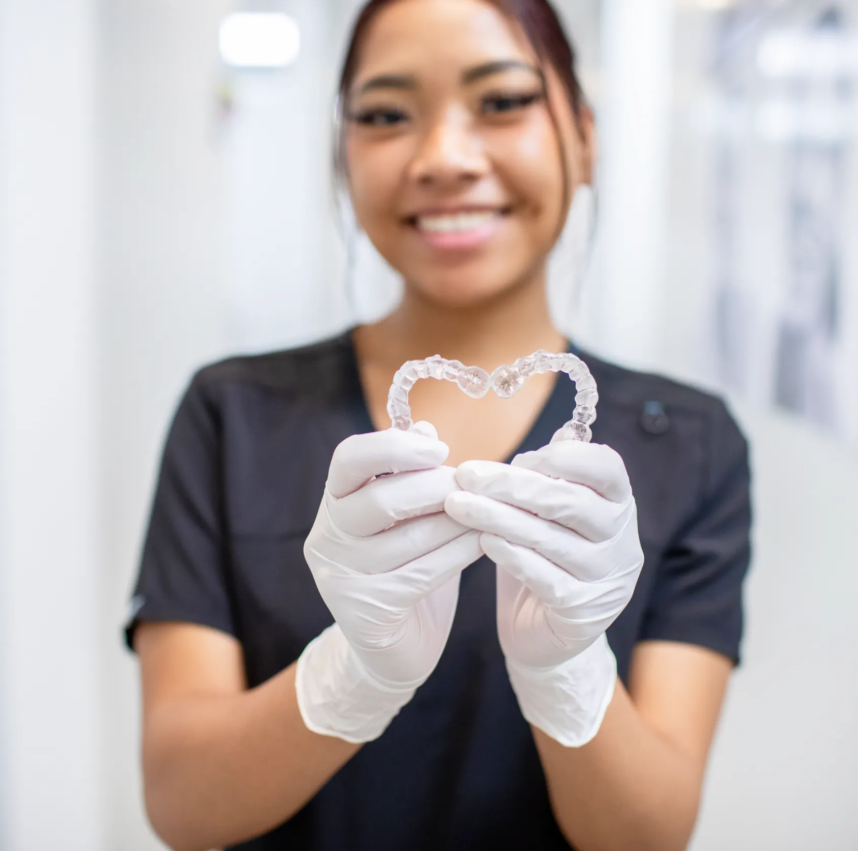 Person wearing gloves holds a pair of dental aligners shaped like a heart, smiling in the background.