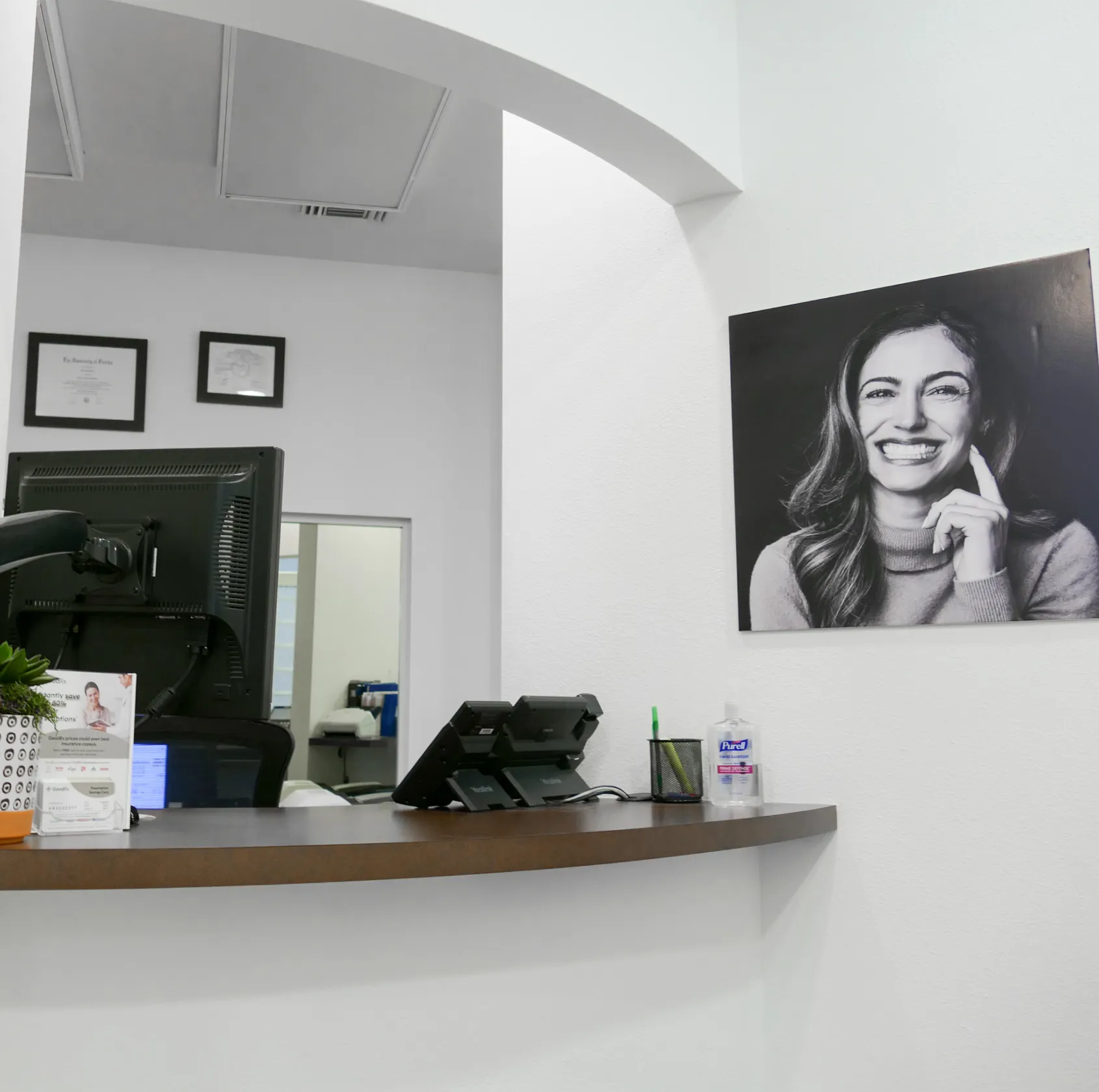 Reception desk with a computer monitor and hand sanitizer, under a large smiling portrait on the wall.