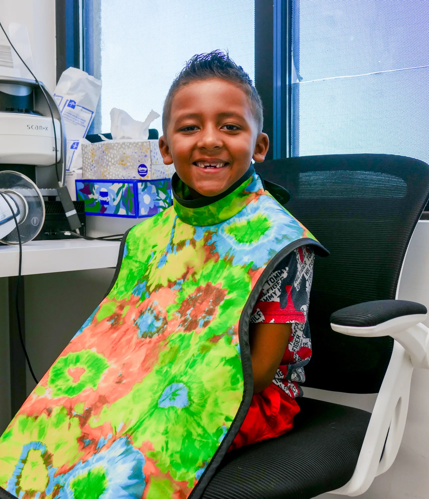 Smiling child wearing a colorful protective cape sits in a chair, possibly at a dental or medical office.