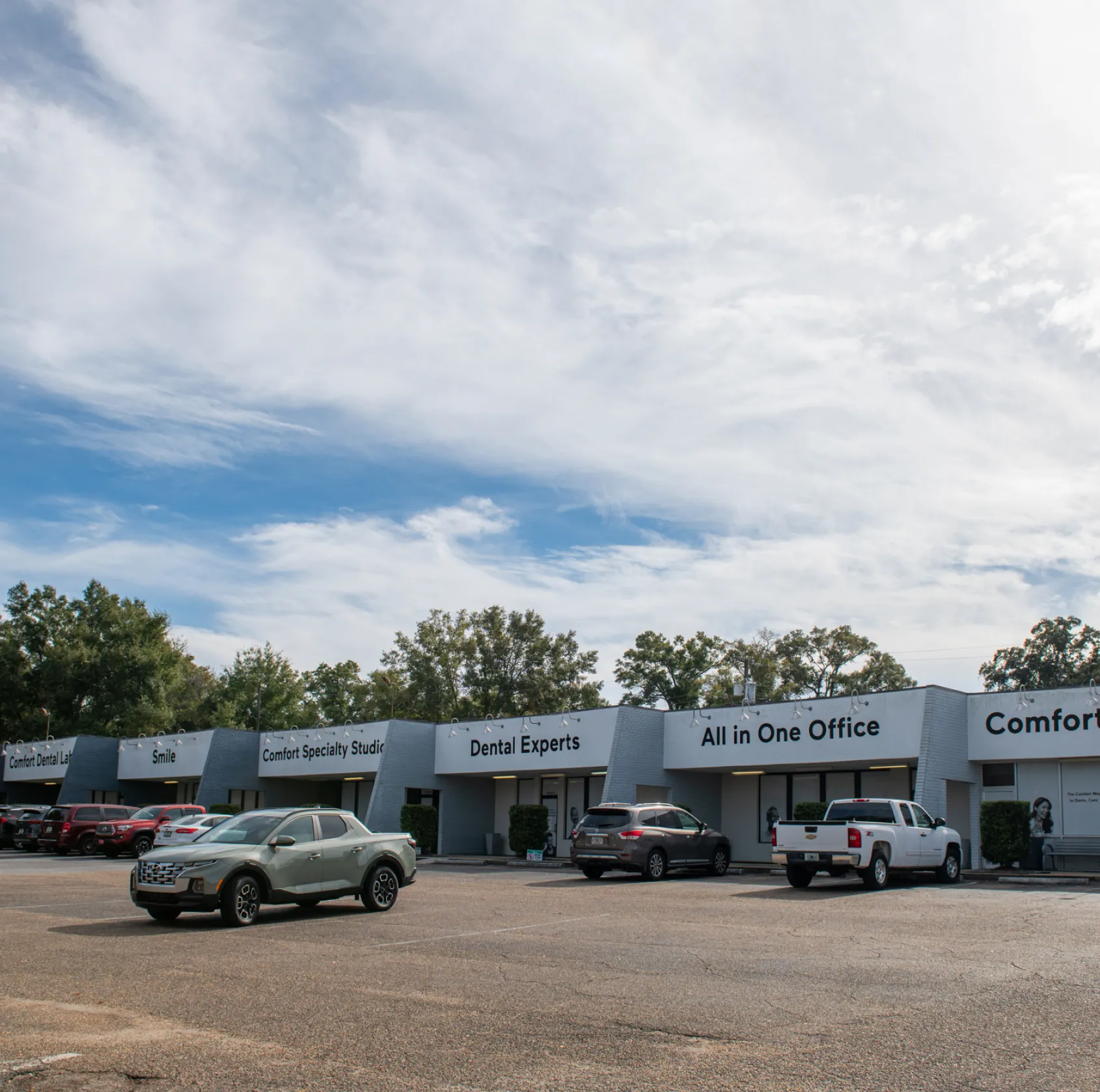 A person with curly blonde hair smiles while wearing a dark shirt against a leafy background.