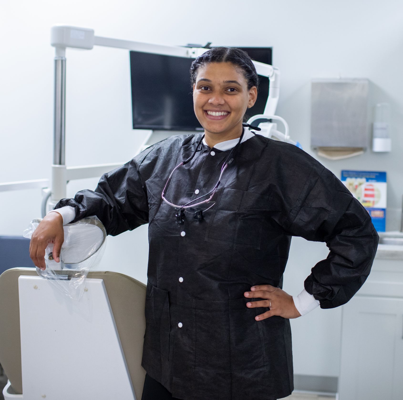 A woman in a dental office wearing a black protective coat stands smiling beside a dental chair.
