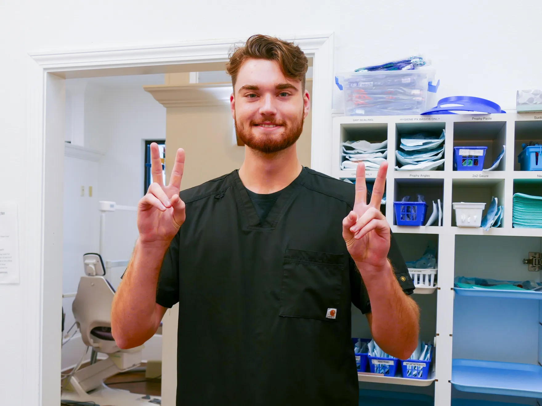 A woman wearing glasses and a blue outfit smiles while standing in an office with computers.