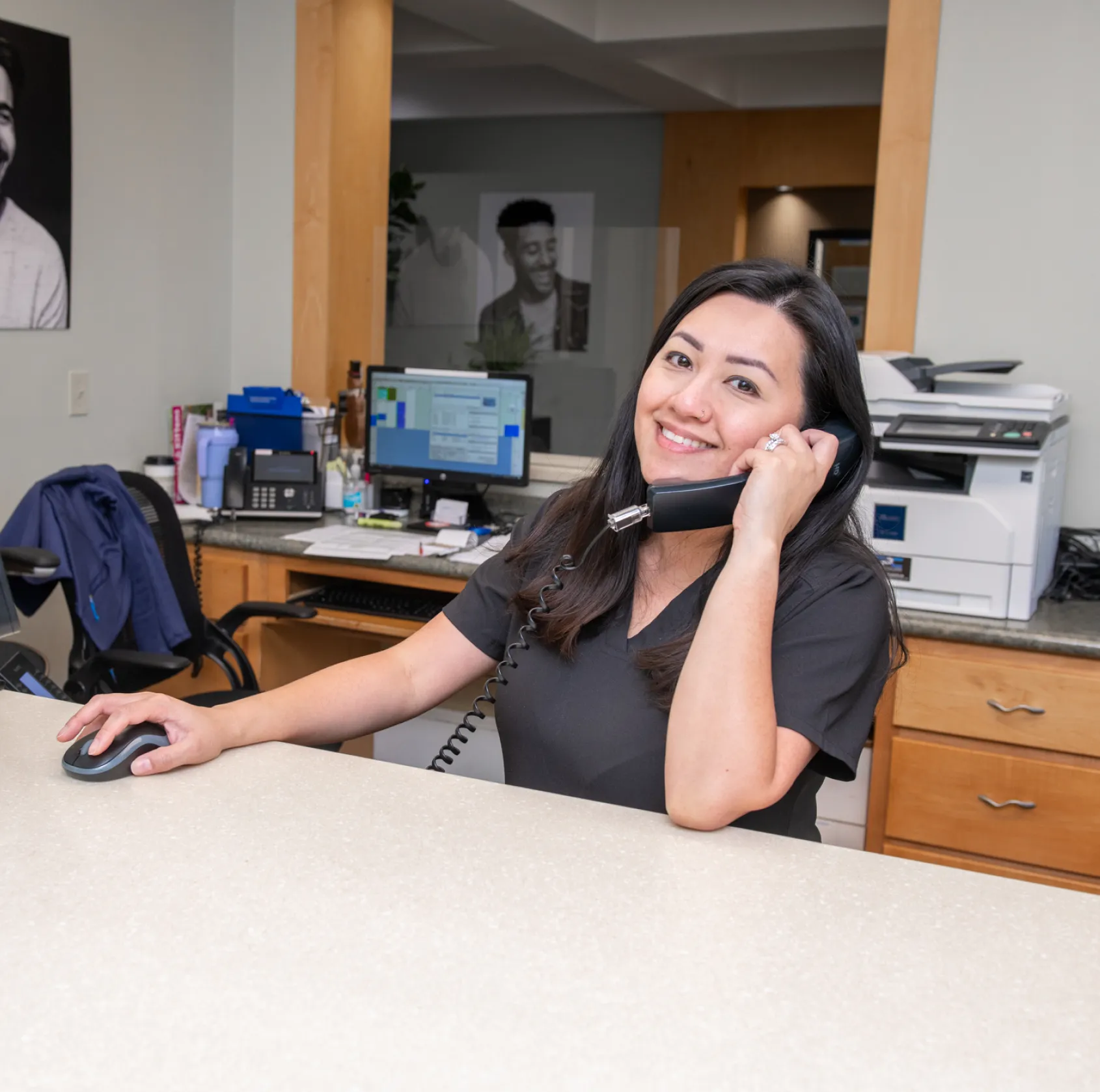 A woman, sitting at a desk, smiles while talking on the phone and using a computer mouse.