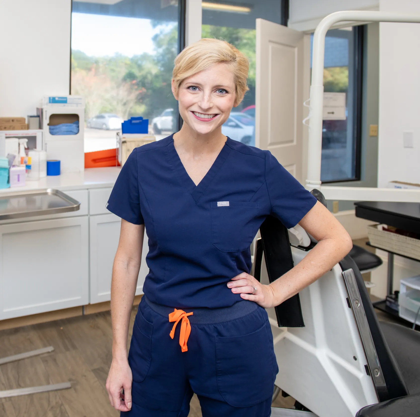 A person in navy scrubs stands smiling in a dental office.