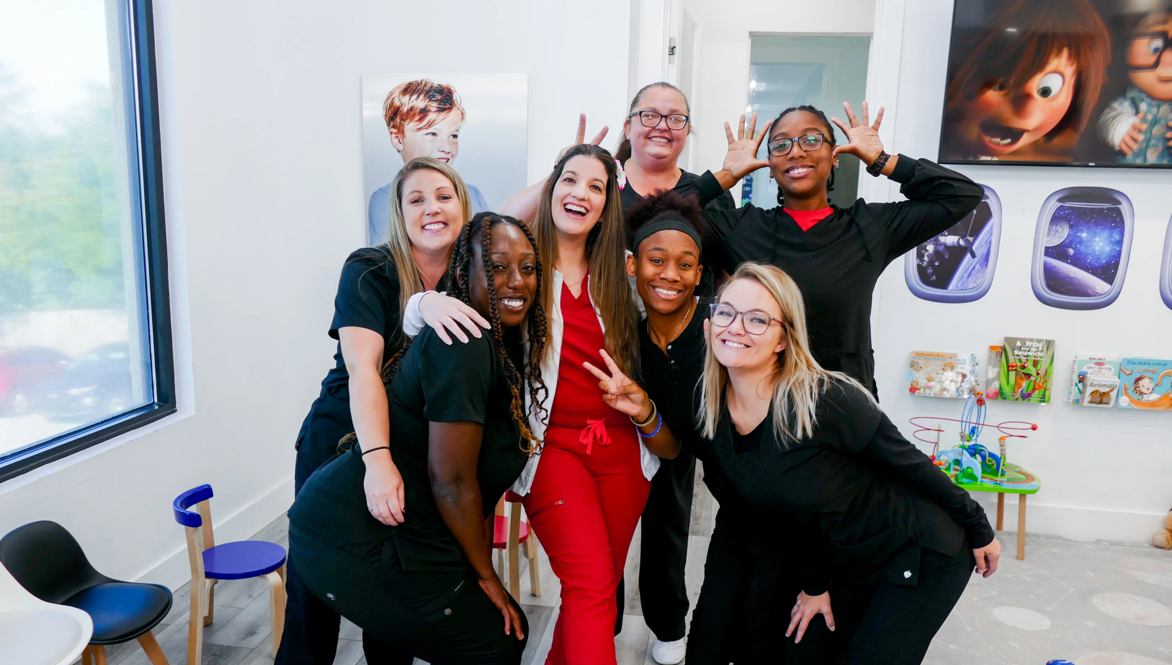 Group of seven smiling people posing together indoors, some making peace signs and playful gestures.