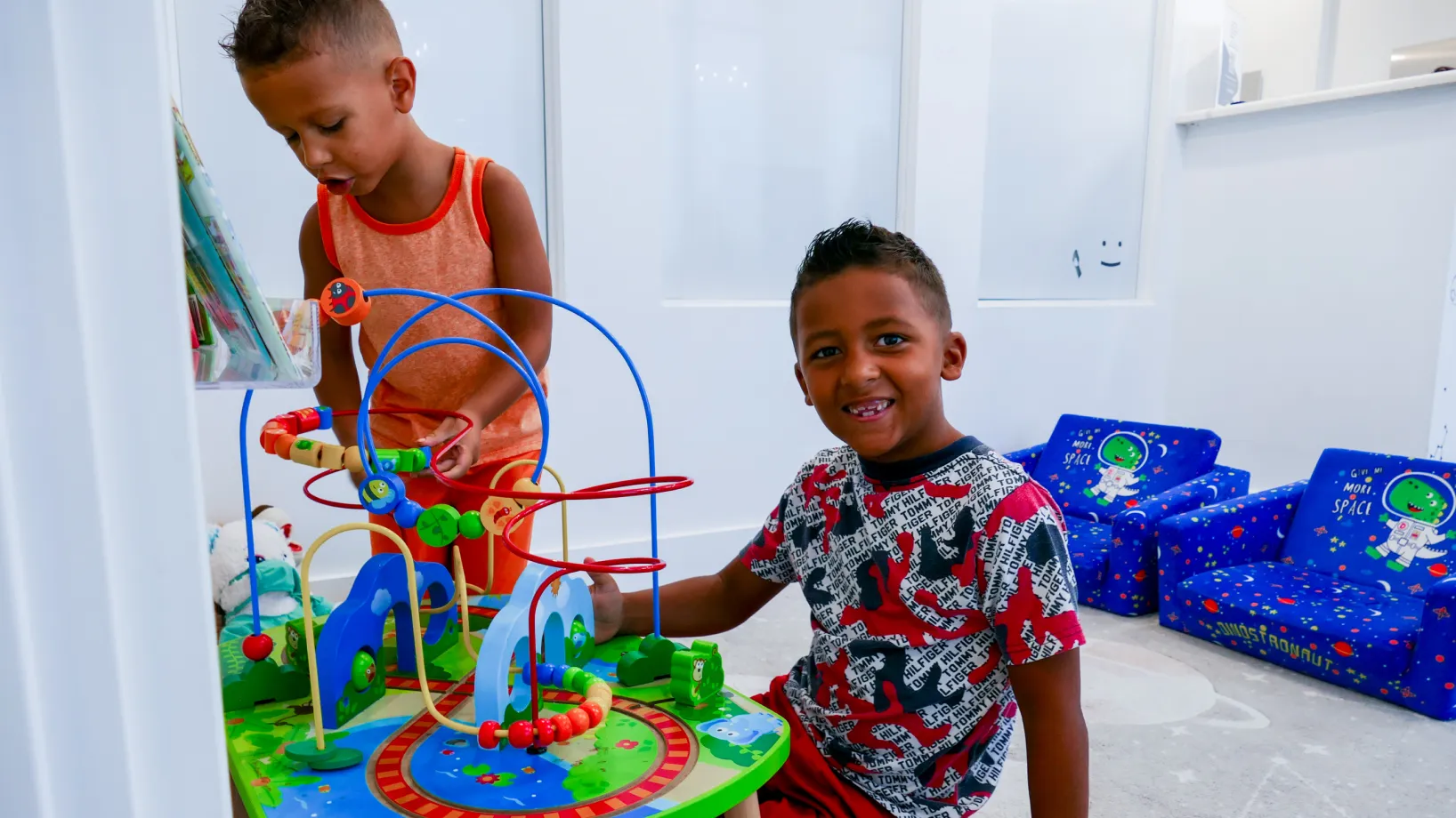 Two boys sitting on blue couches, smiling at the camera, one holding a green tablet.