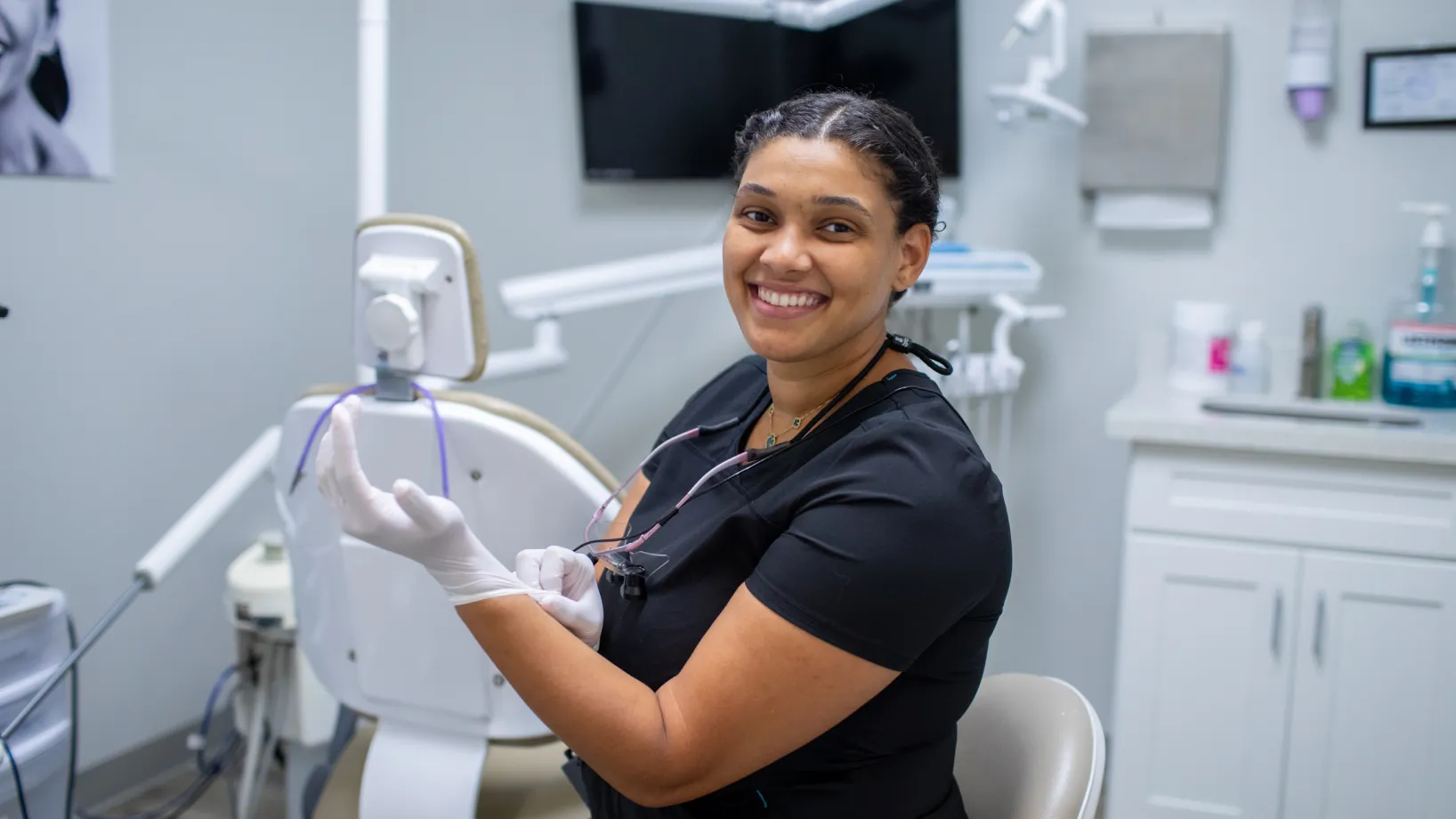 An empty dental chair in a clinic room with medical equipment and cabinets nearby.
