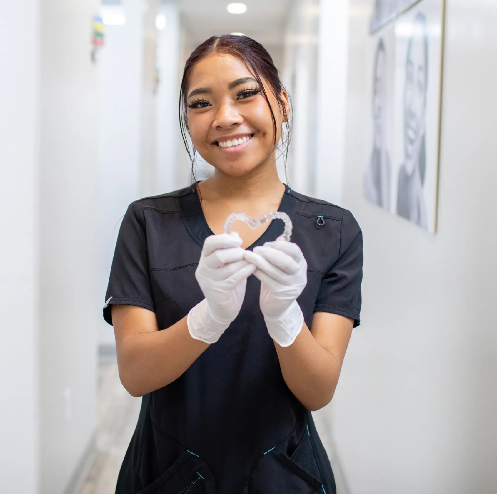 A smiling person in scrubs and gloves holds clear dental aligners in both hands.