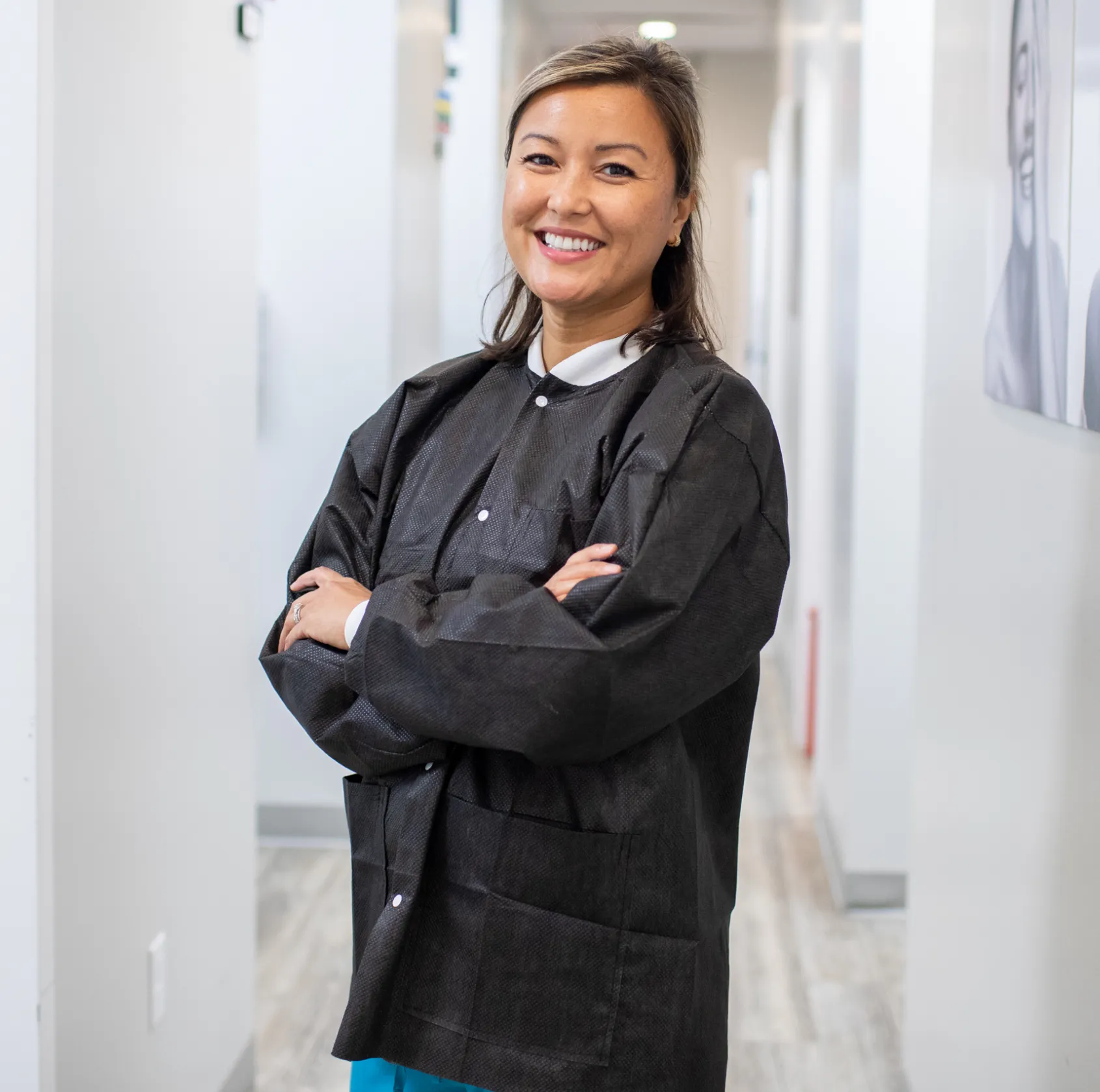 Smiling woman in a black jacket stands with arms crossed in a hallway.
