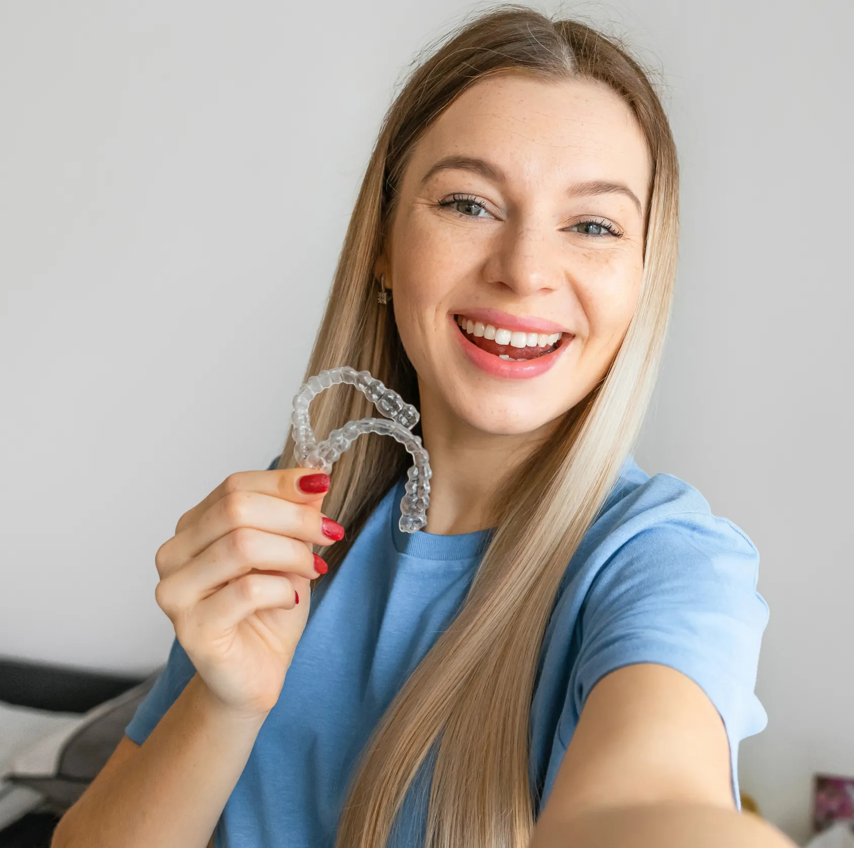 A smiling woman holds a clear dental aligner, showing her teeth, wearing a blue shirt.