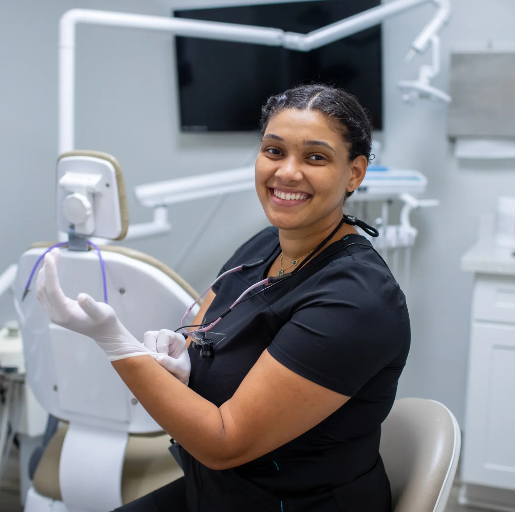 A dental professional smiles while putting on gloves, sitting in a dental clinic.