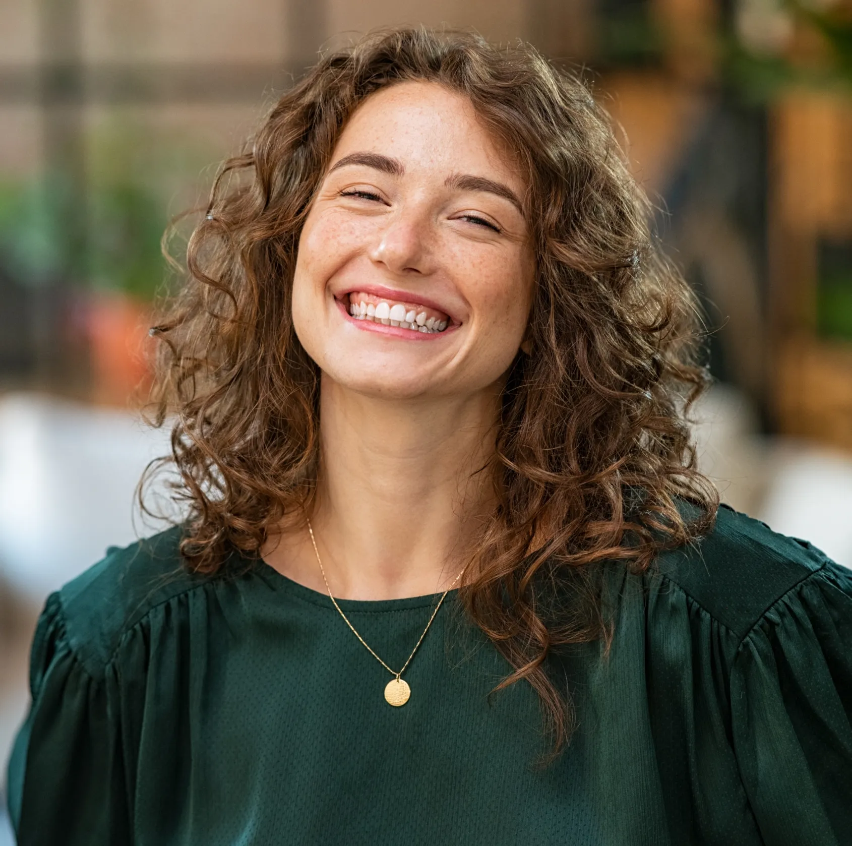 A person with curly hair smiles broadly, wearing a green blouse and a gold necklace.