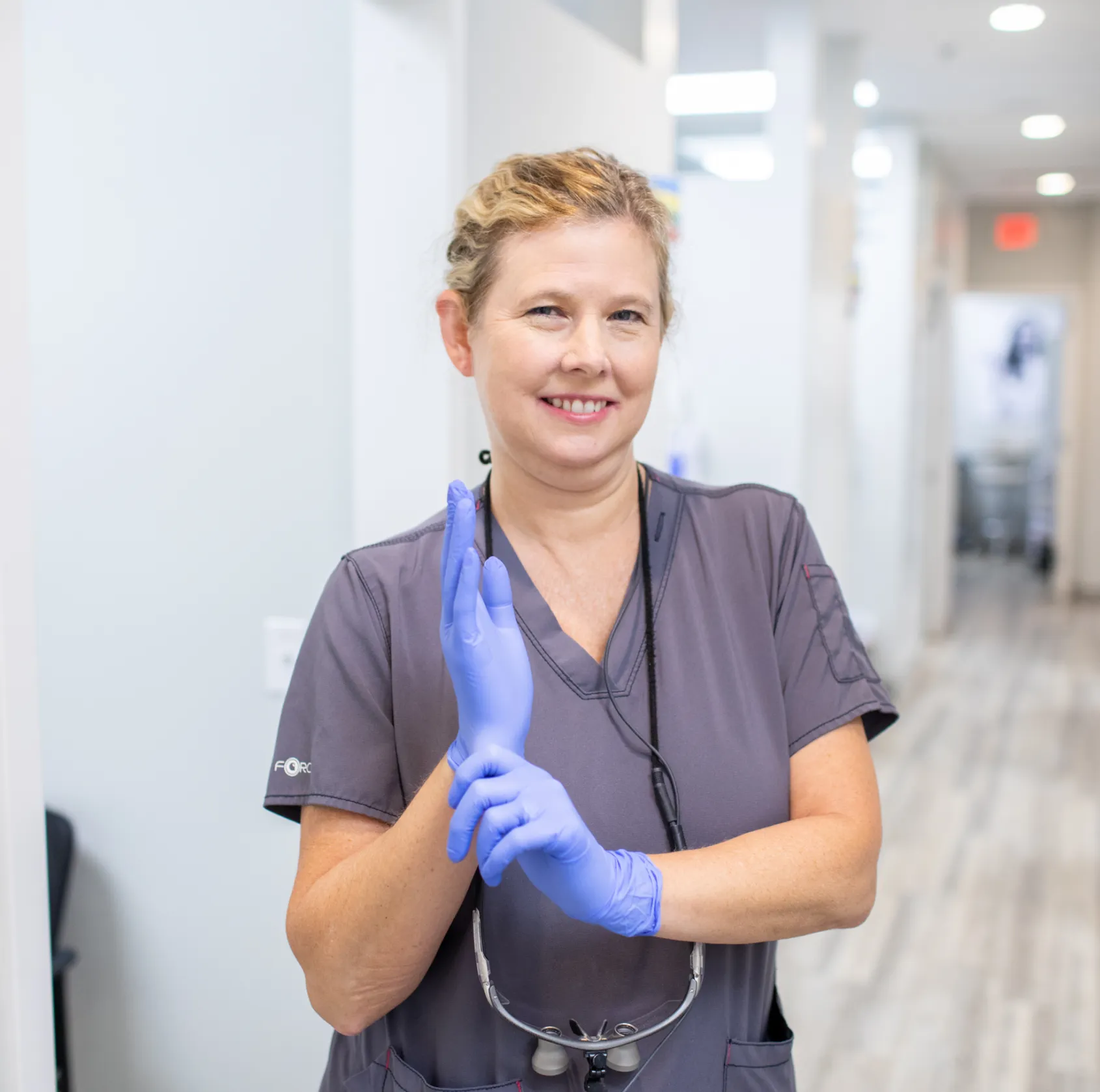 A woman in scrubs smiles while putting on blue gloves in a medical setting.