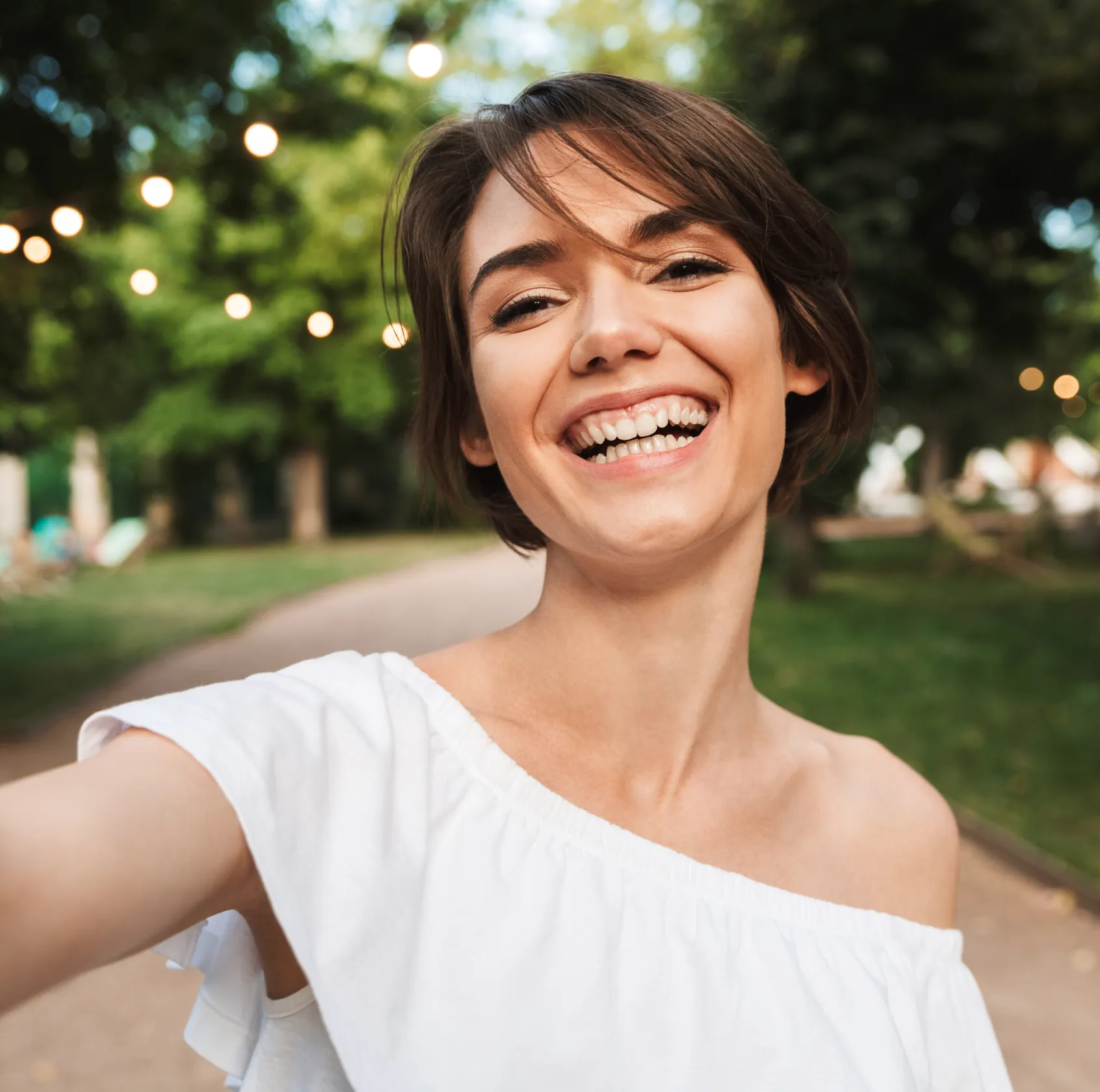 A person smiling broadly while taking a selfie in a park with string lights above.