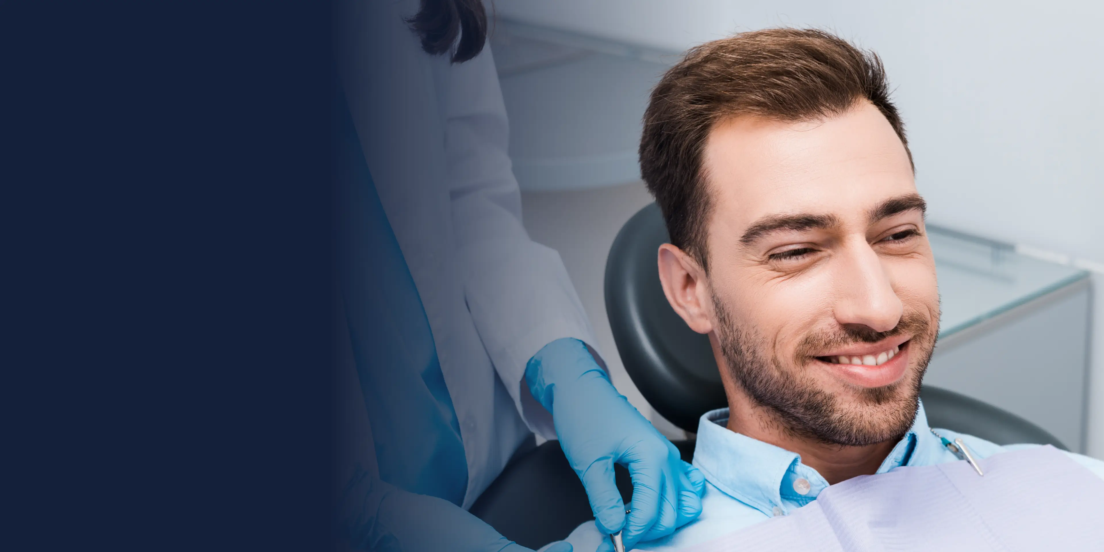 A man sits in a dental chair, smiling, as a dentist in gloves prepares to examine him.
