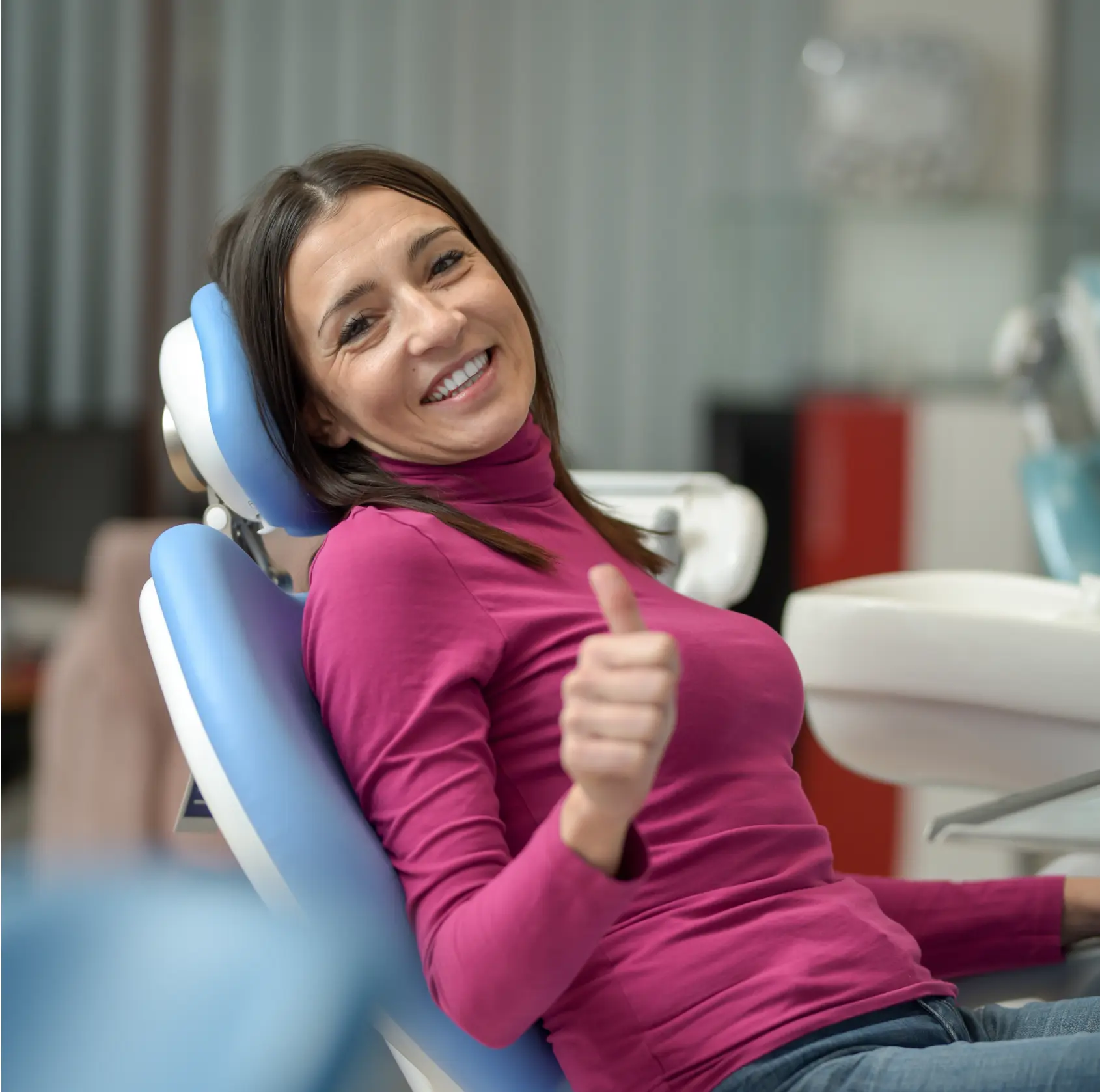 A woman in a pink shirt smiles and gives a thumbs up while sitting in a dental chair.