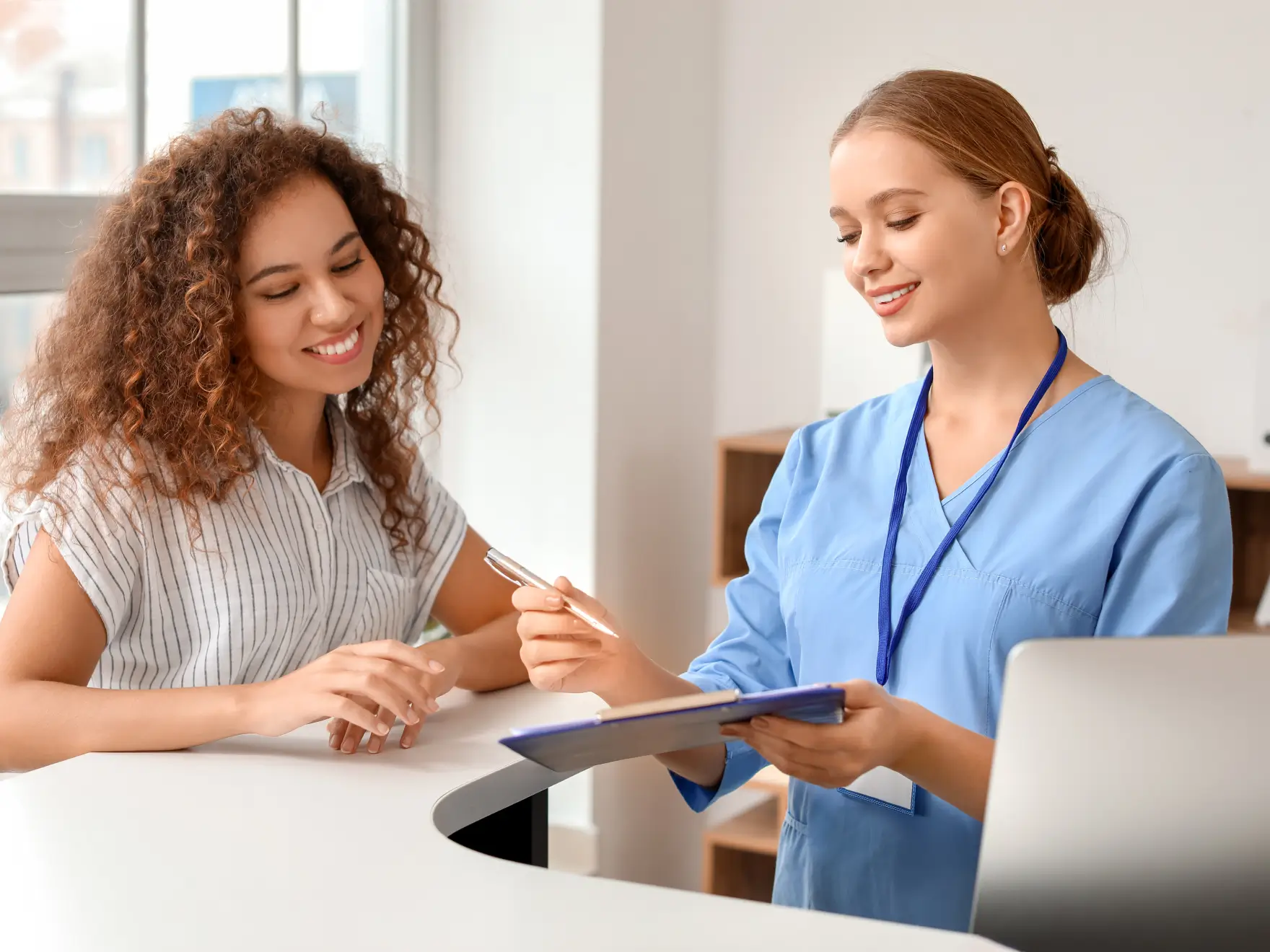 A nurse in blue scrubs helps a young woman at a reception desk.