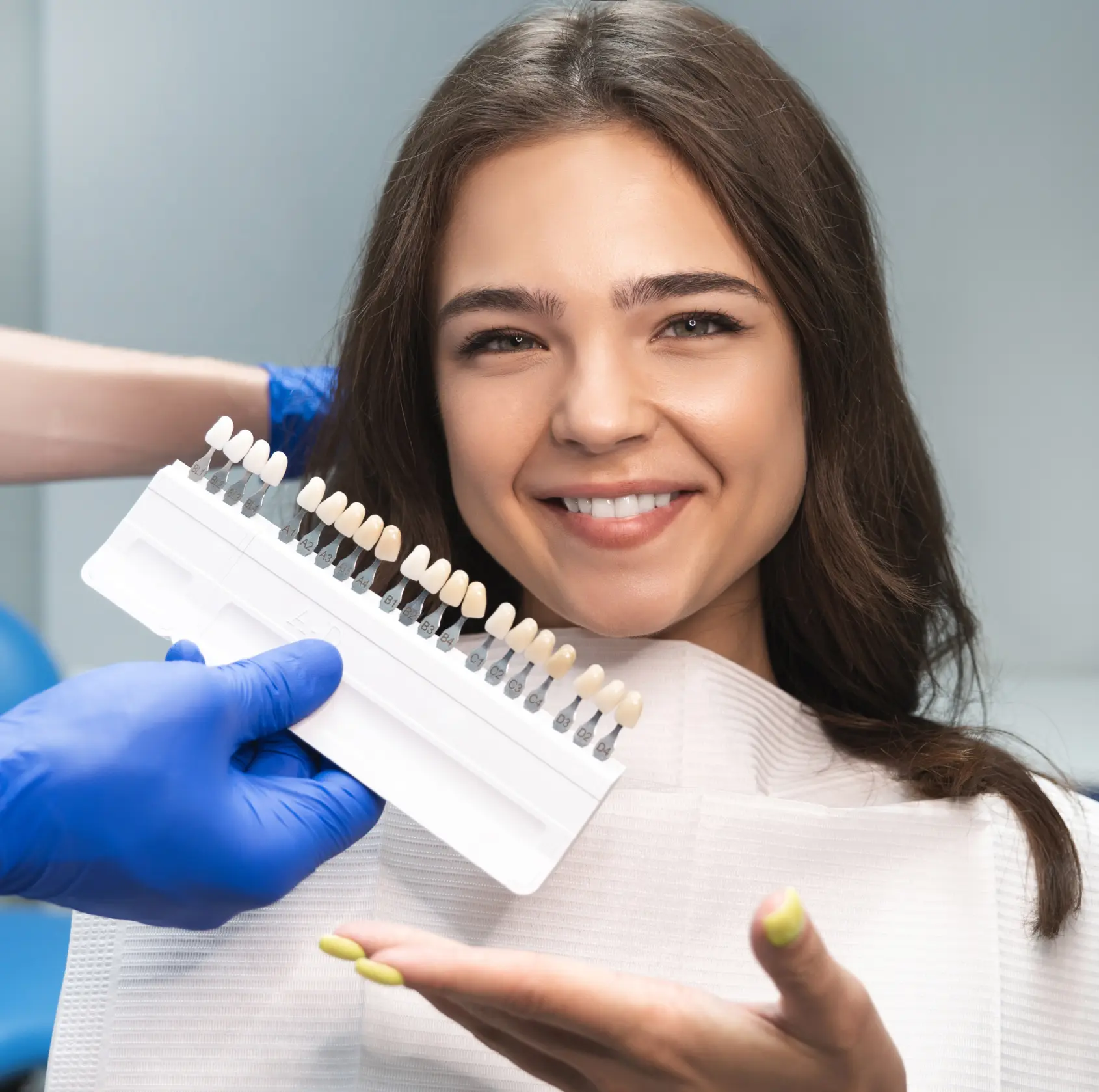 A woman smiling while a dental professional holds a tooth color chart near her face.