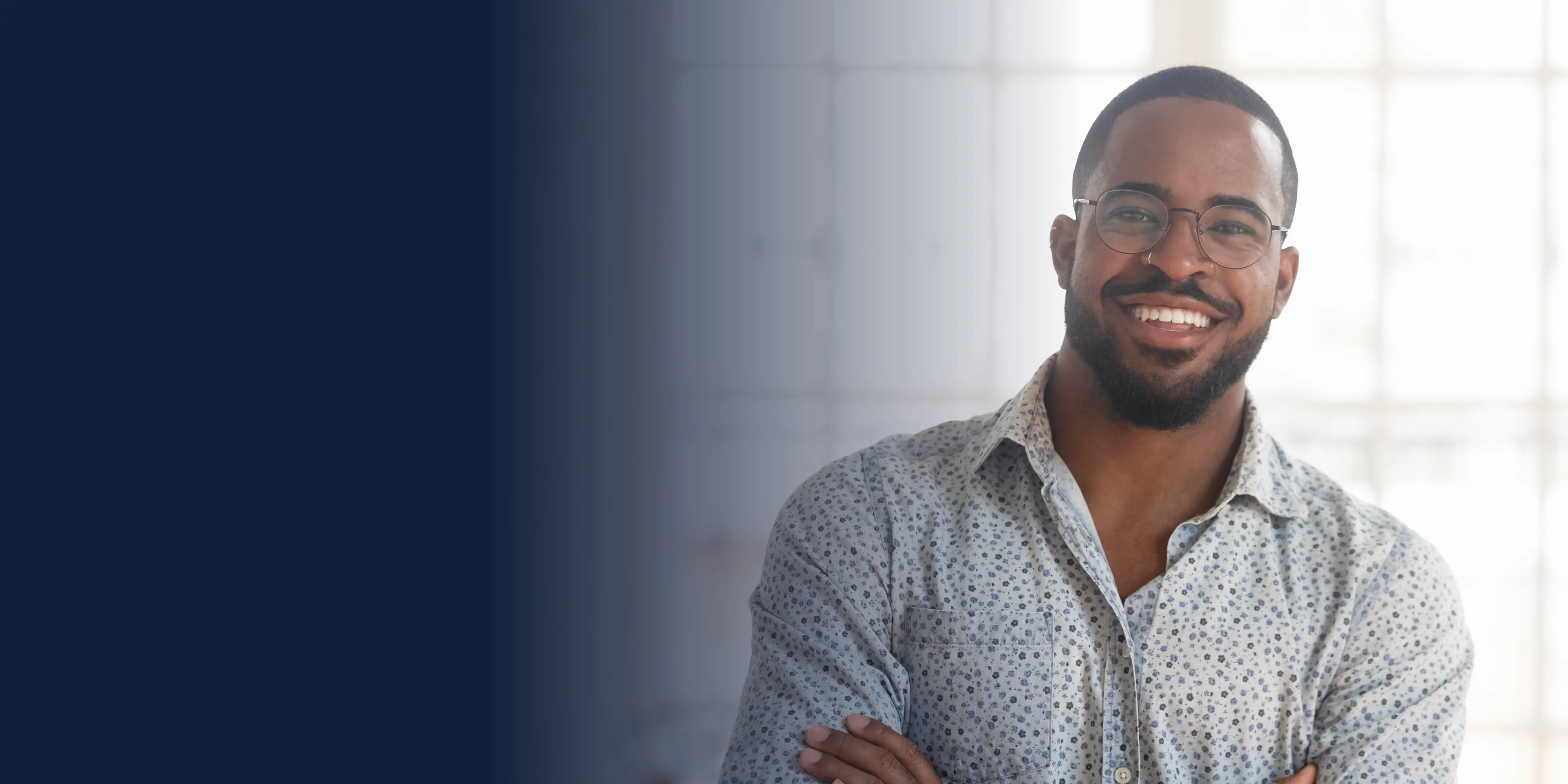 Man with glasses smiling, wearing a light-patterned shirt, standing with arms crossed indoors.