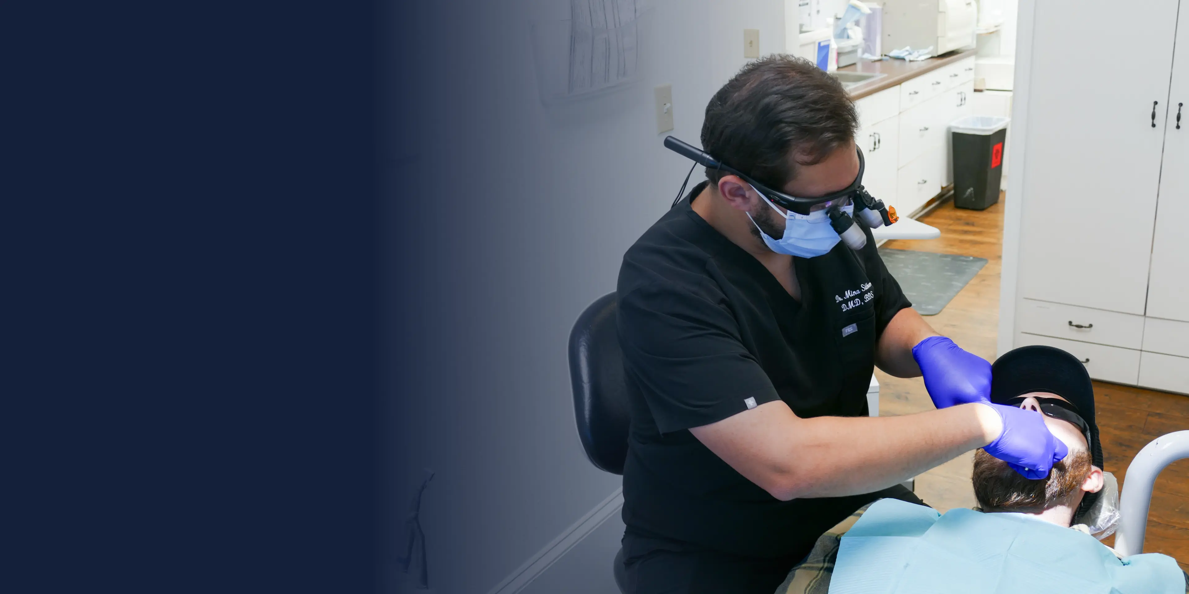 A dentist wearing magnifying glasses and gloves performs a procedure on a reclining patient.