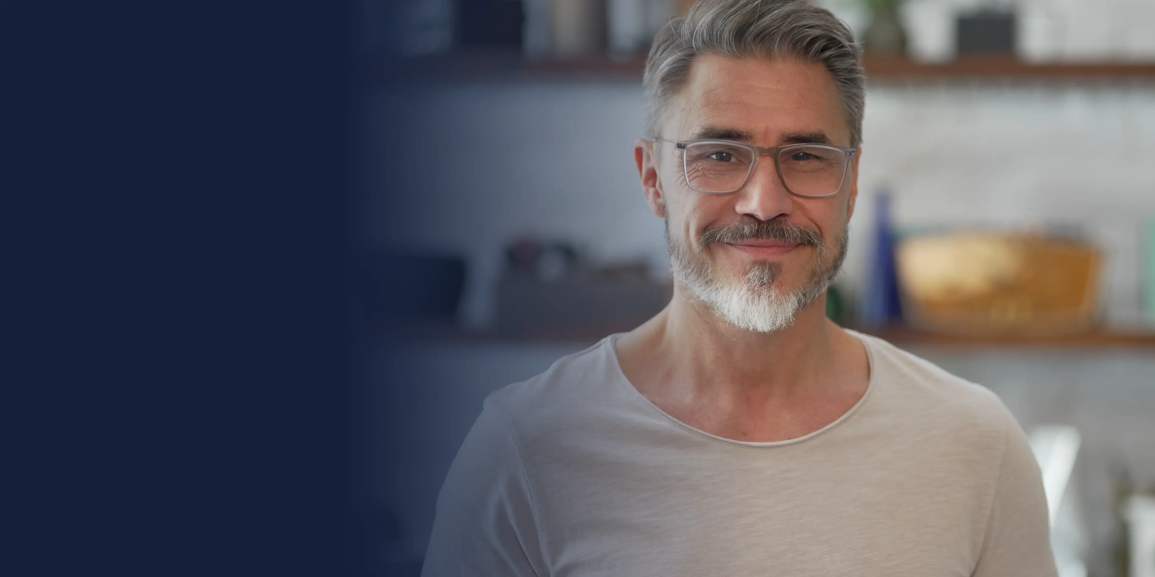 Man with gray hair and glasses smiling while standing indoors wearing a light-colored shirt.