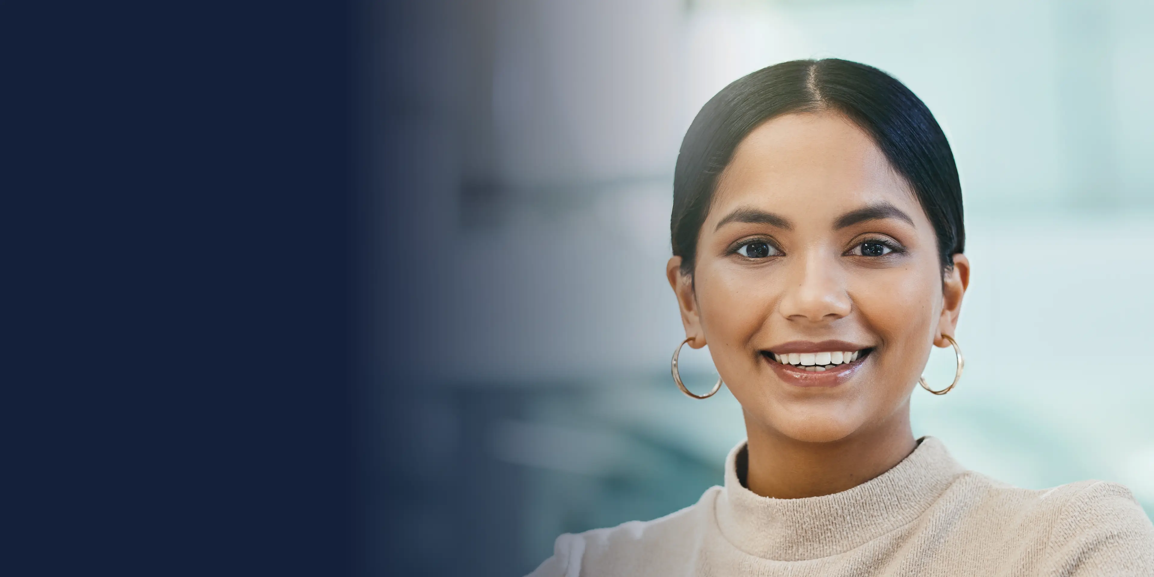 Smiling person with dark hair, wearing hoop earrings and a beige turtleneck, against a neutral background.
