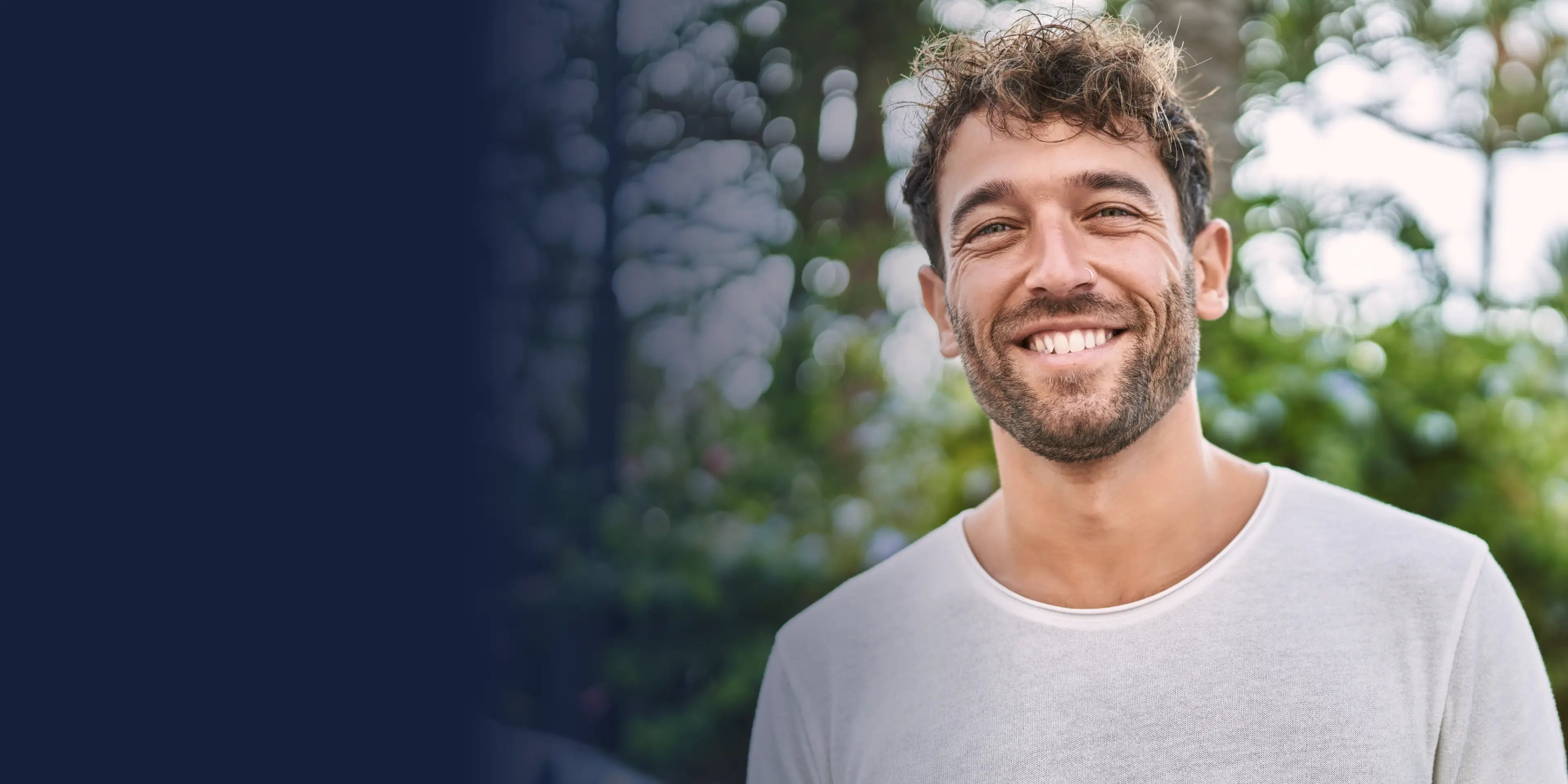 A man with curly hair smiles warmly, standing outside in front of green foliage.