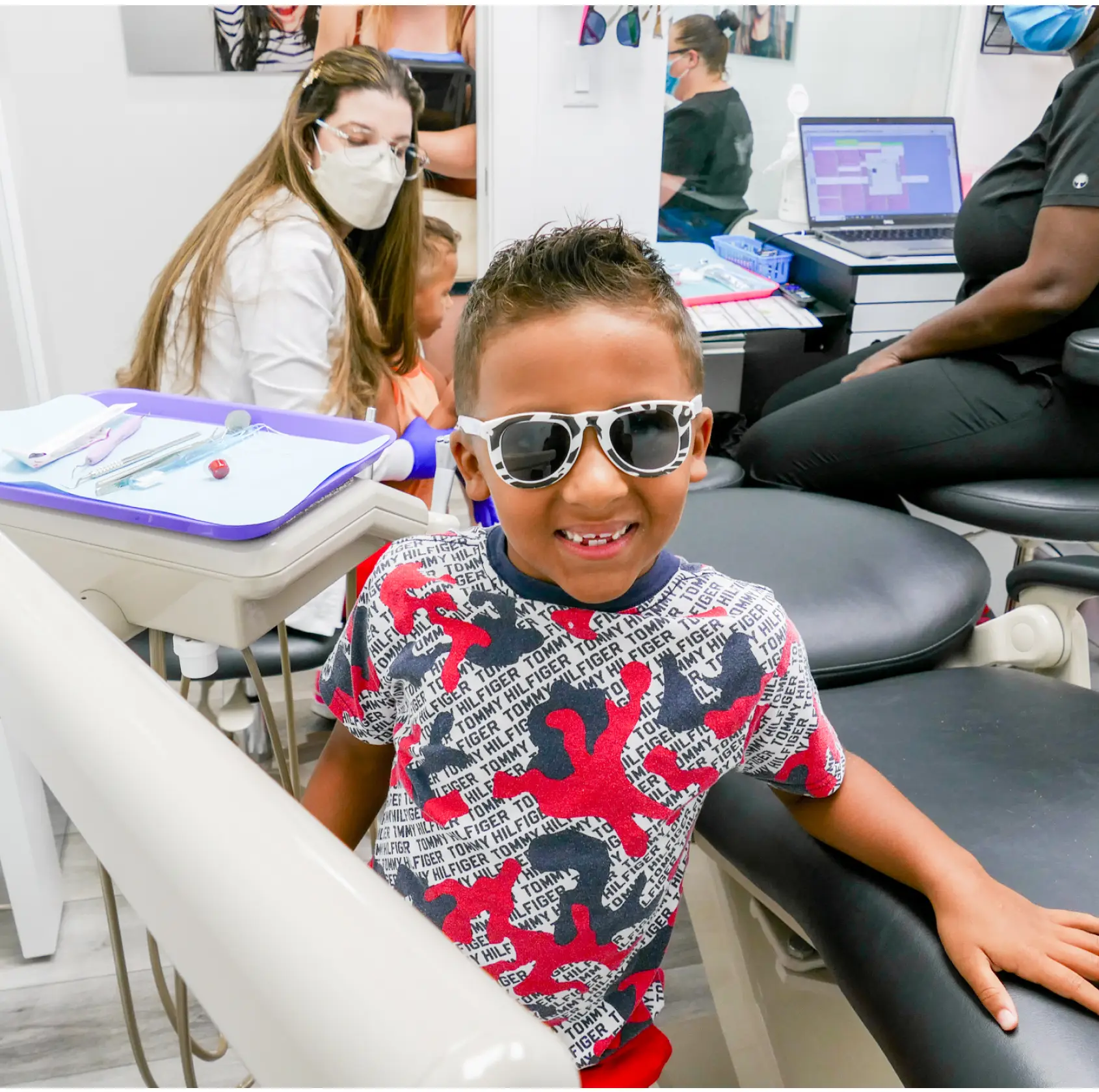 Young boy smiling and wearing sunglasses at a dental office, with dental staff nearby.