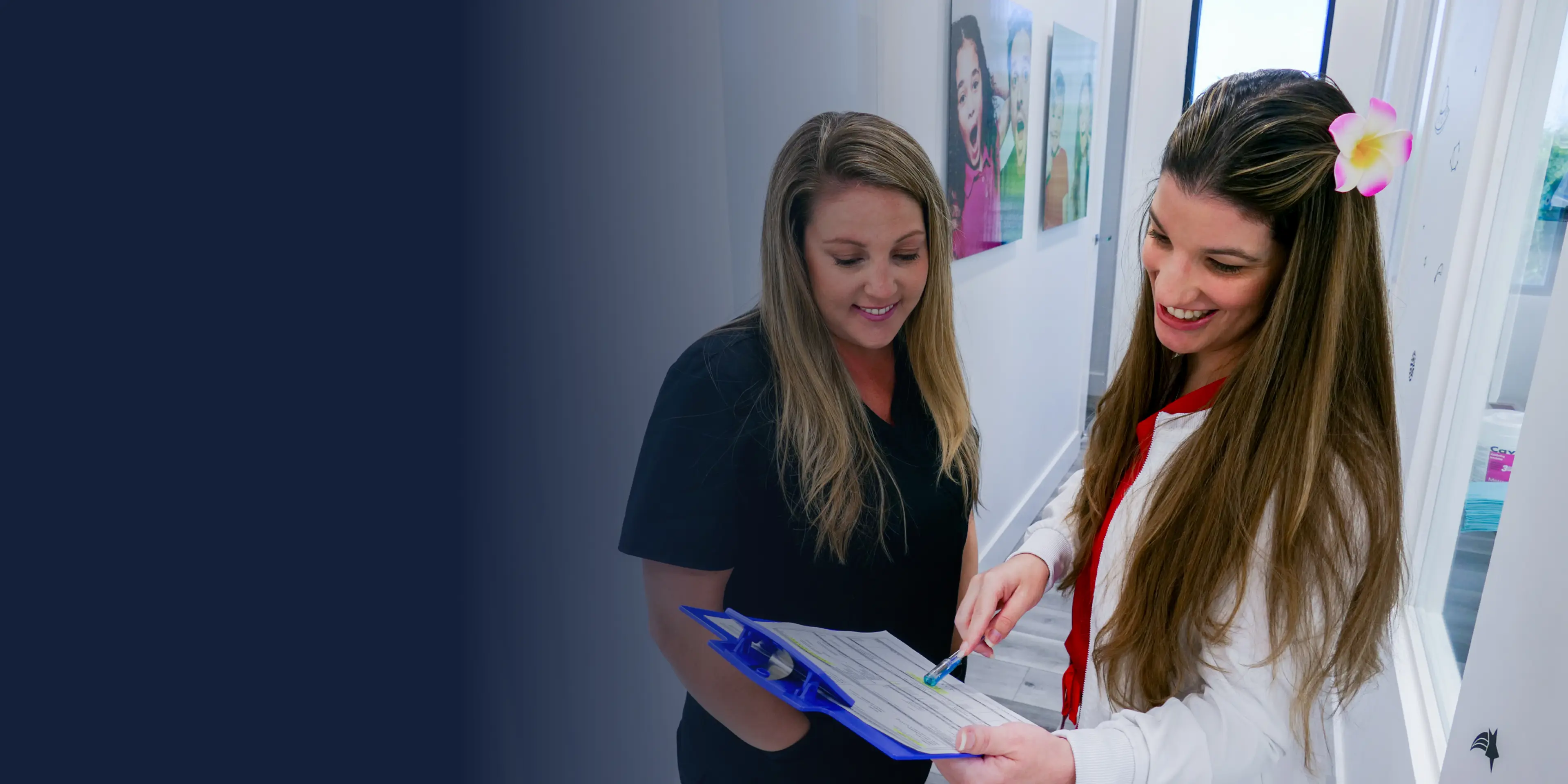 Two women stand in a hallway, discussing a document on a blue clipboard.