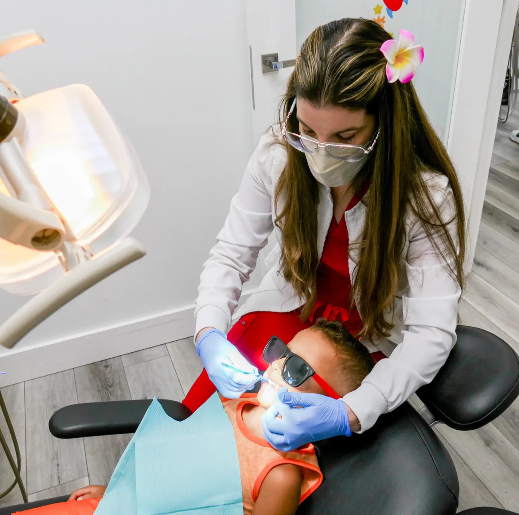 Dentist wearing gloves examines a child's teeth under a bright light.
