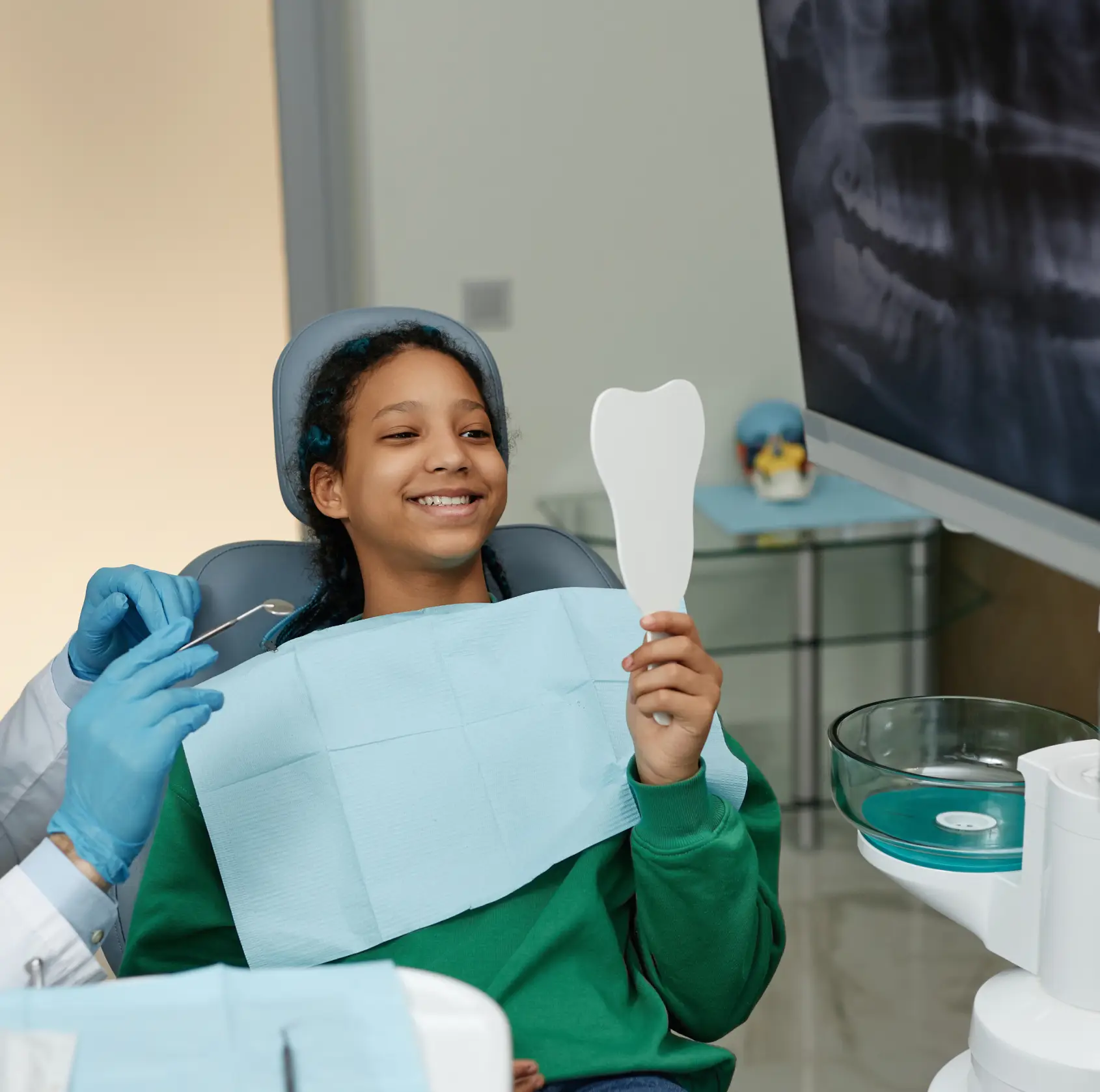 A person in a dental chair smiles while looking at their teeth in a handheld mirror.