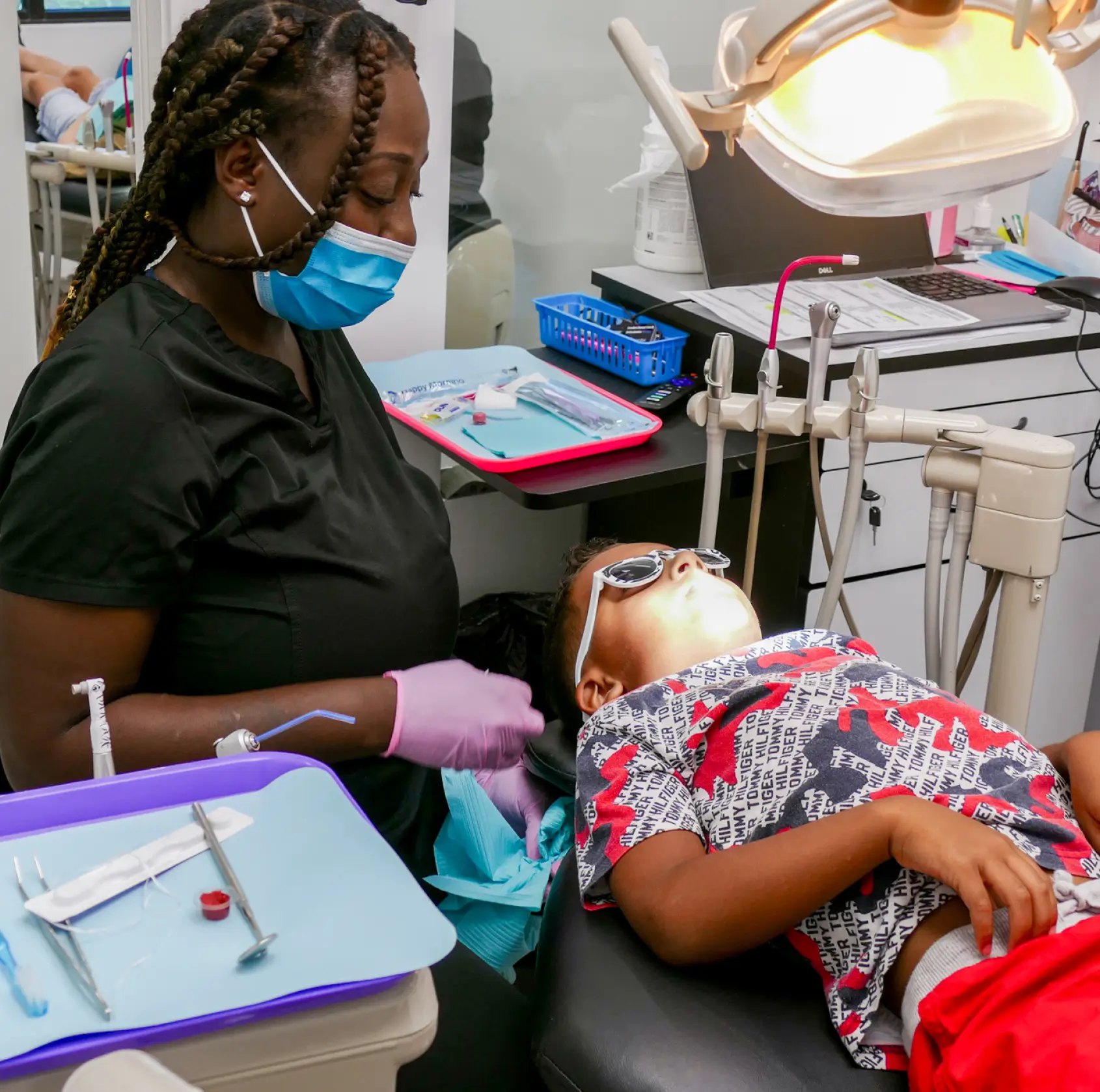 A dental hygienist wearing a mask and gloves attends to a child lying on a dental chair.