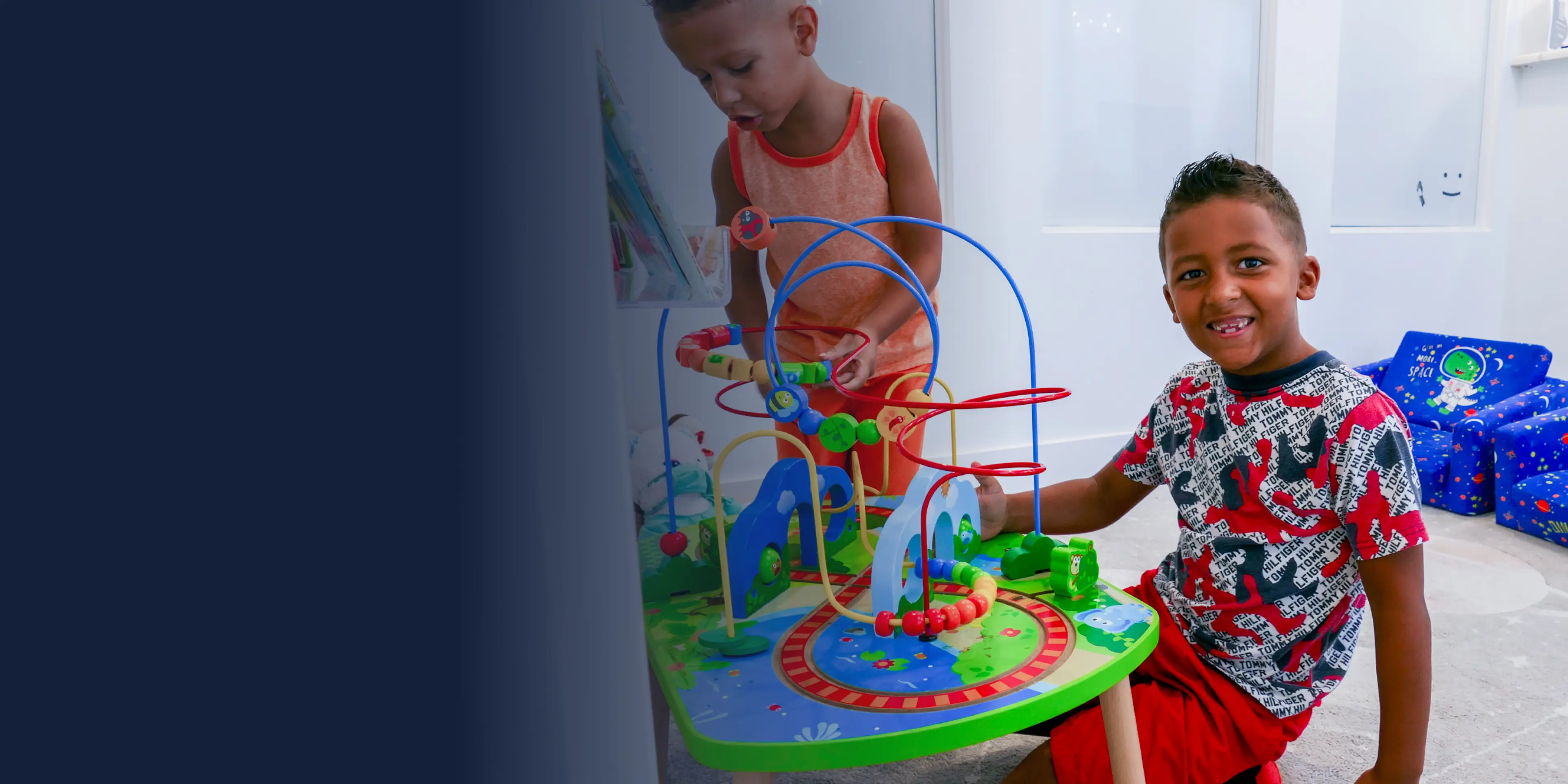 Two children playing with a colorful bead maze on the floor, one smiling and sitting, the other standing.