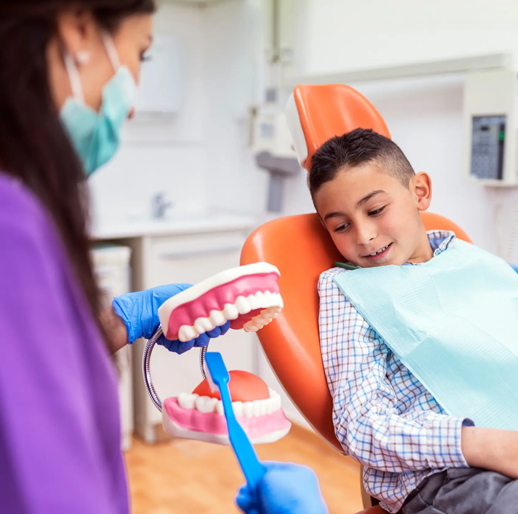 Dentist shows a young boy how to brush teeth using a large model in dental office.