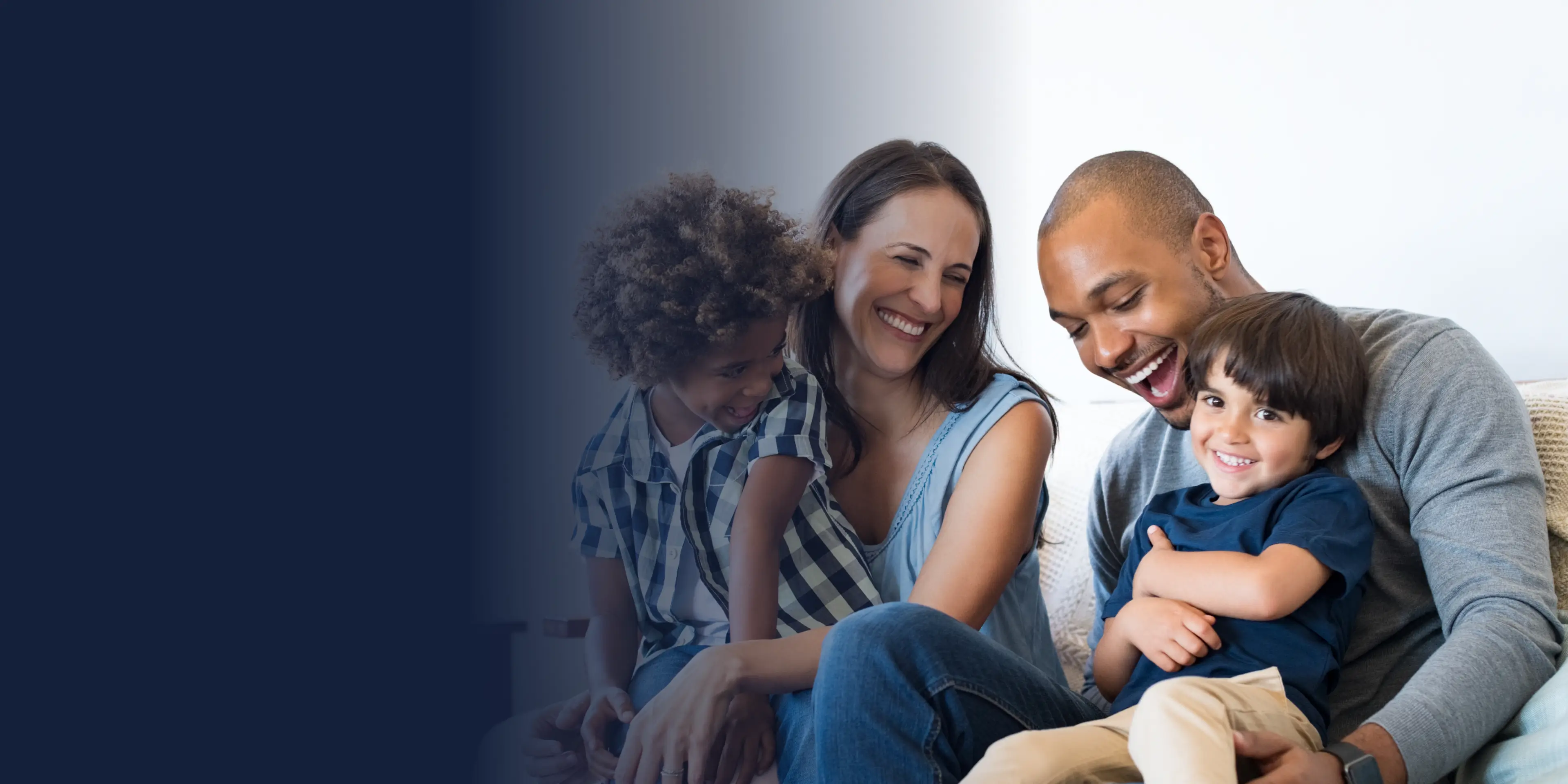 A happy family of four sits closely together on a sofa, smiling and laughing.