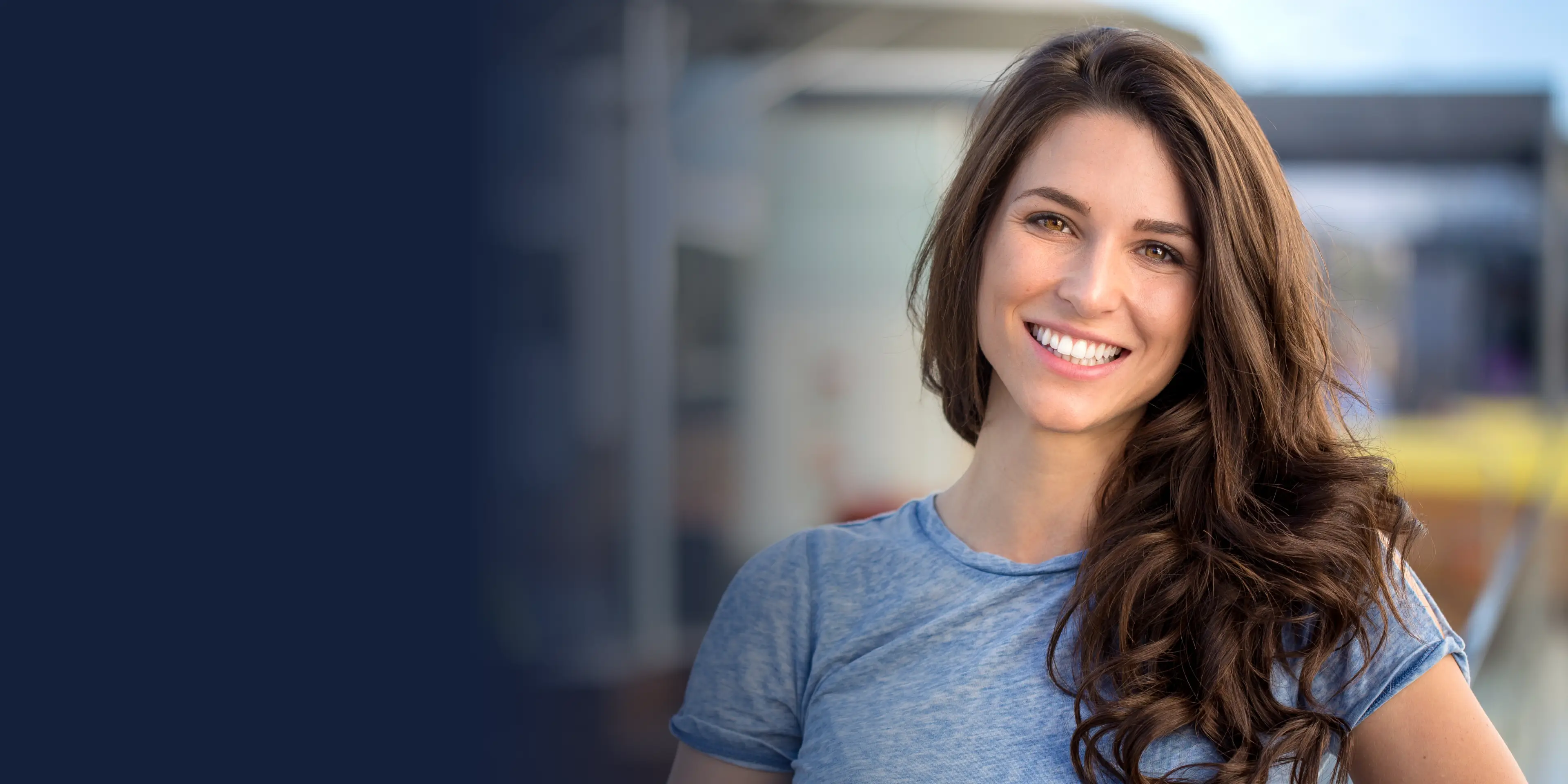 A woman with long brown hair smiles warmly, wearing a light blue shirt.