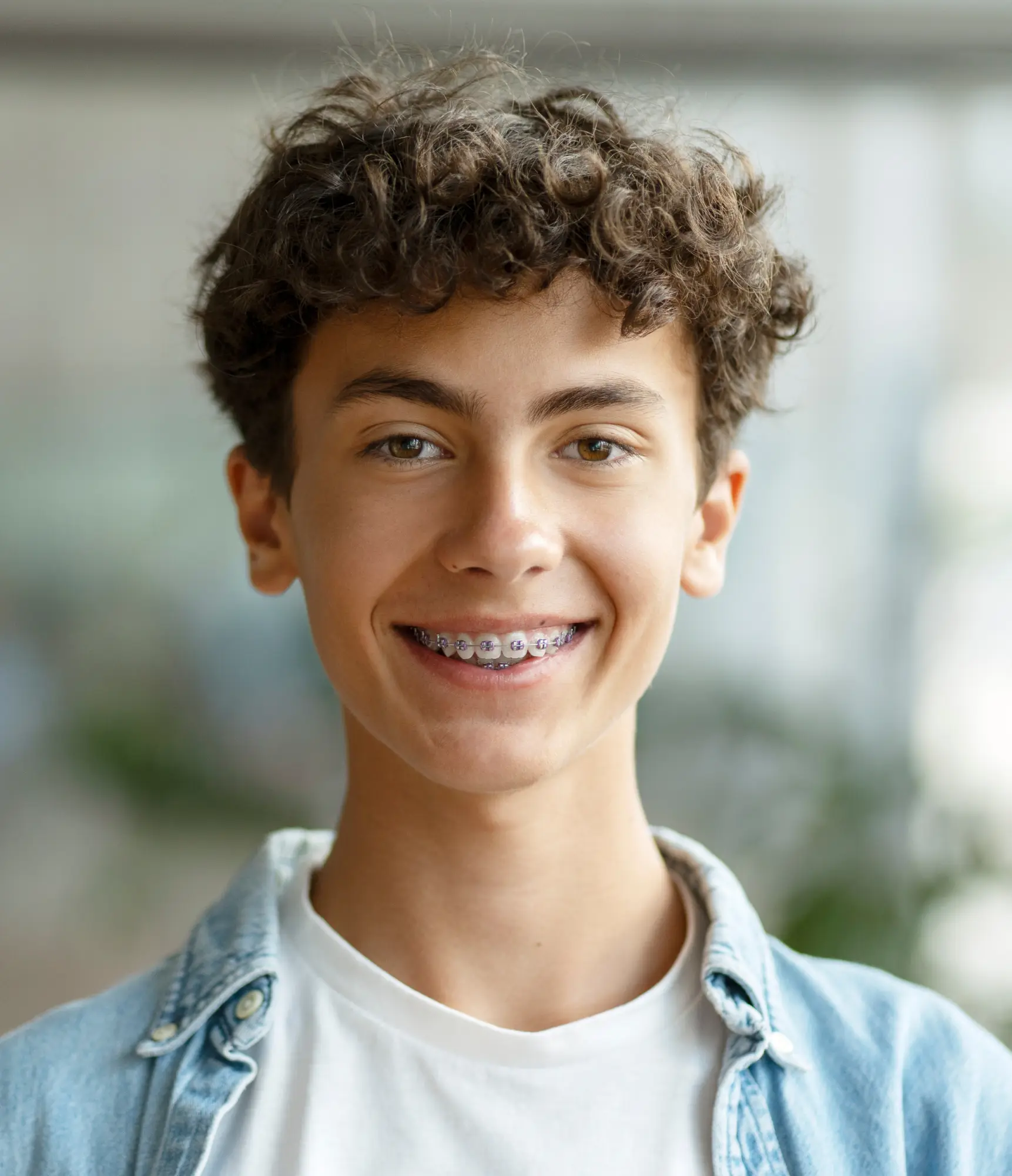 Young person smiling, showing braces, wearing a denim jacket and white shirt.