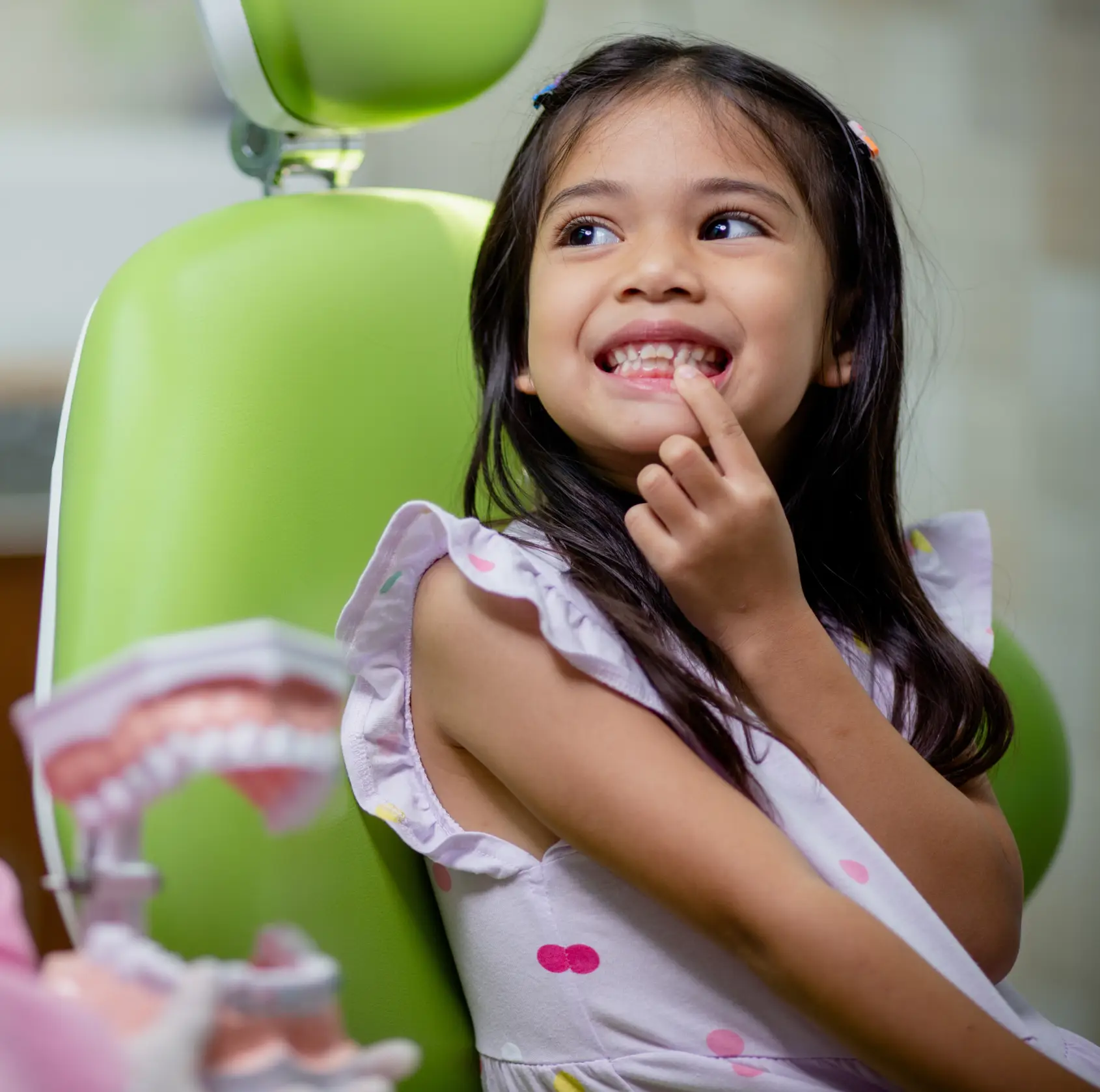 A young girl sits in a dentist's chair, pointing to her teeth and smiling.