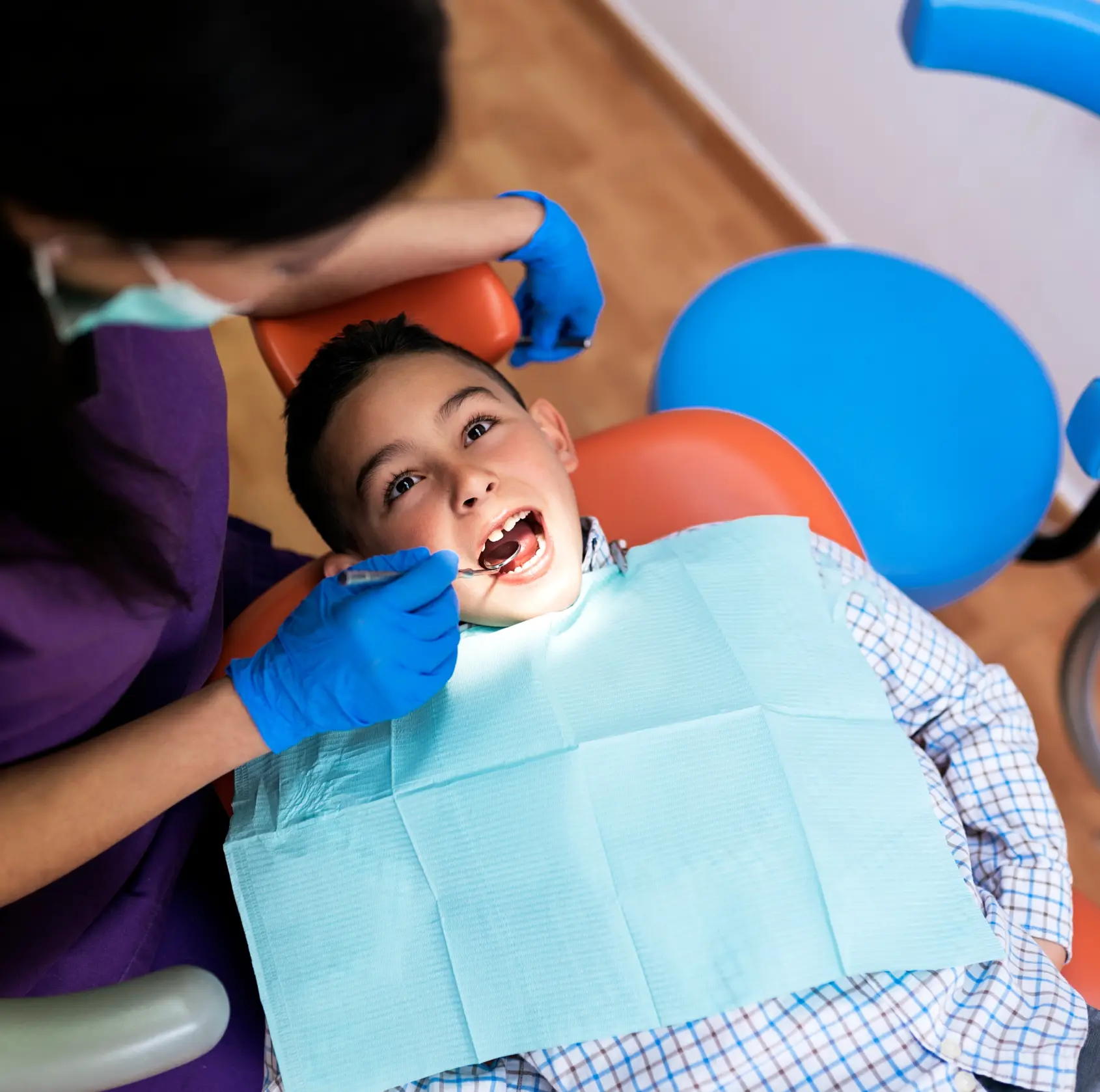 A dentist examines a boy's teeth while he sits in the dental chair with a bib.
