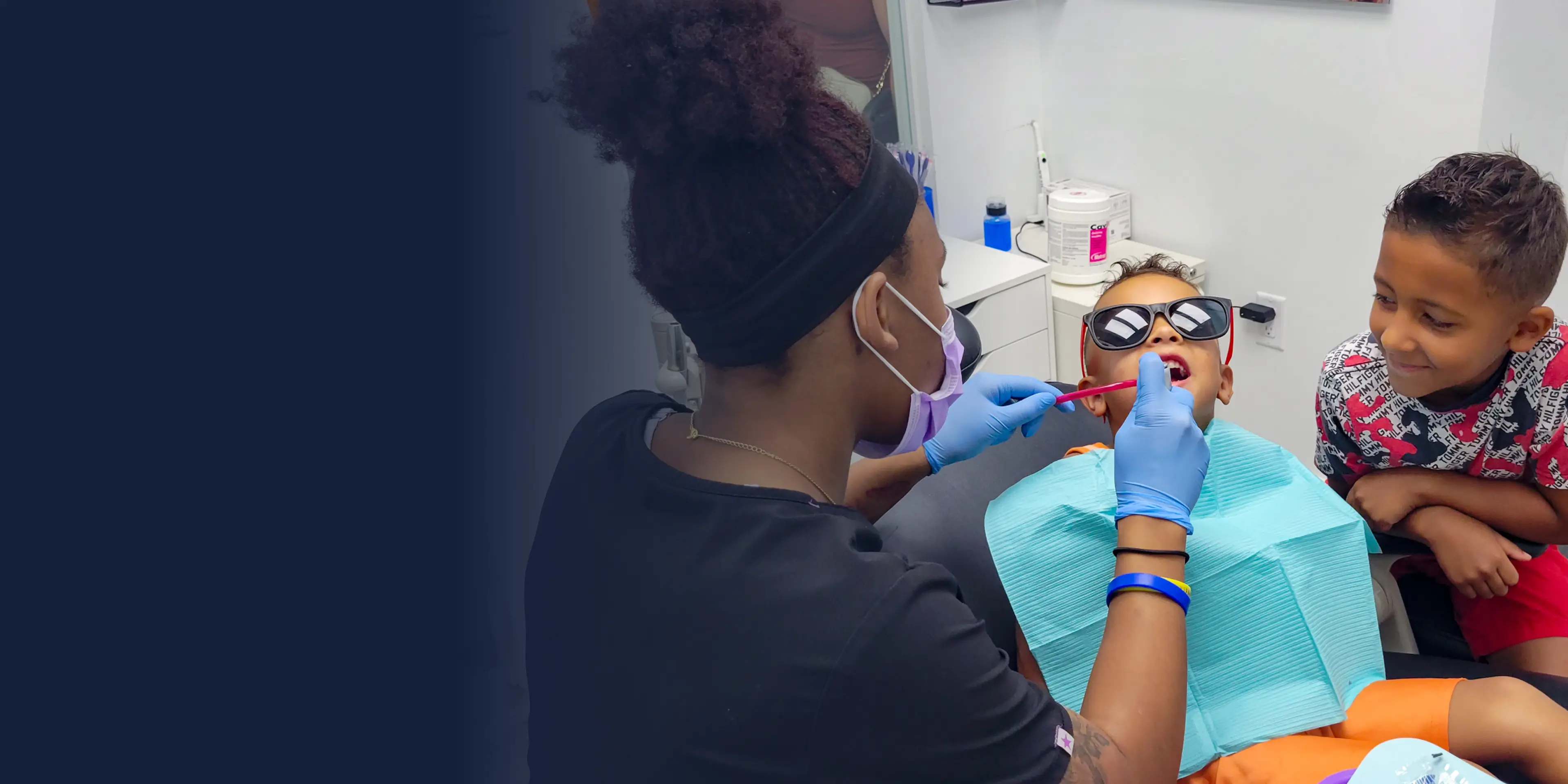 A dental hygienist examines a child's teeth while another child observes nearby.