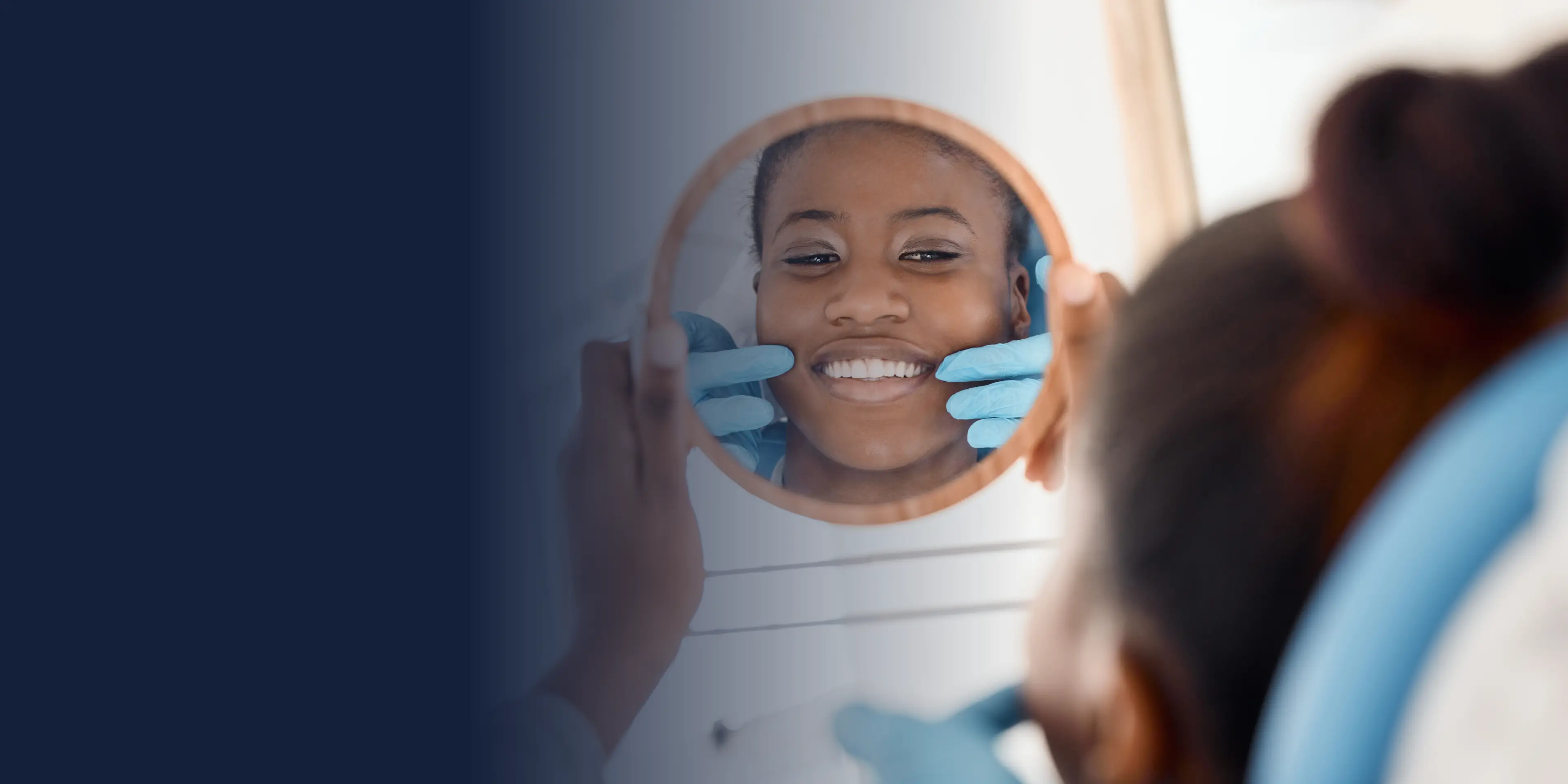 A woman smiles in a small round mirror as someone in gloves checks her teeth.