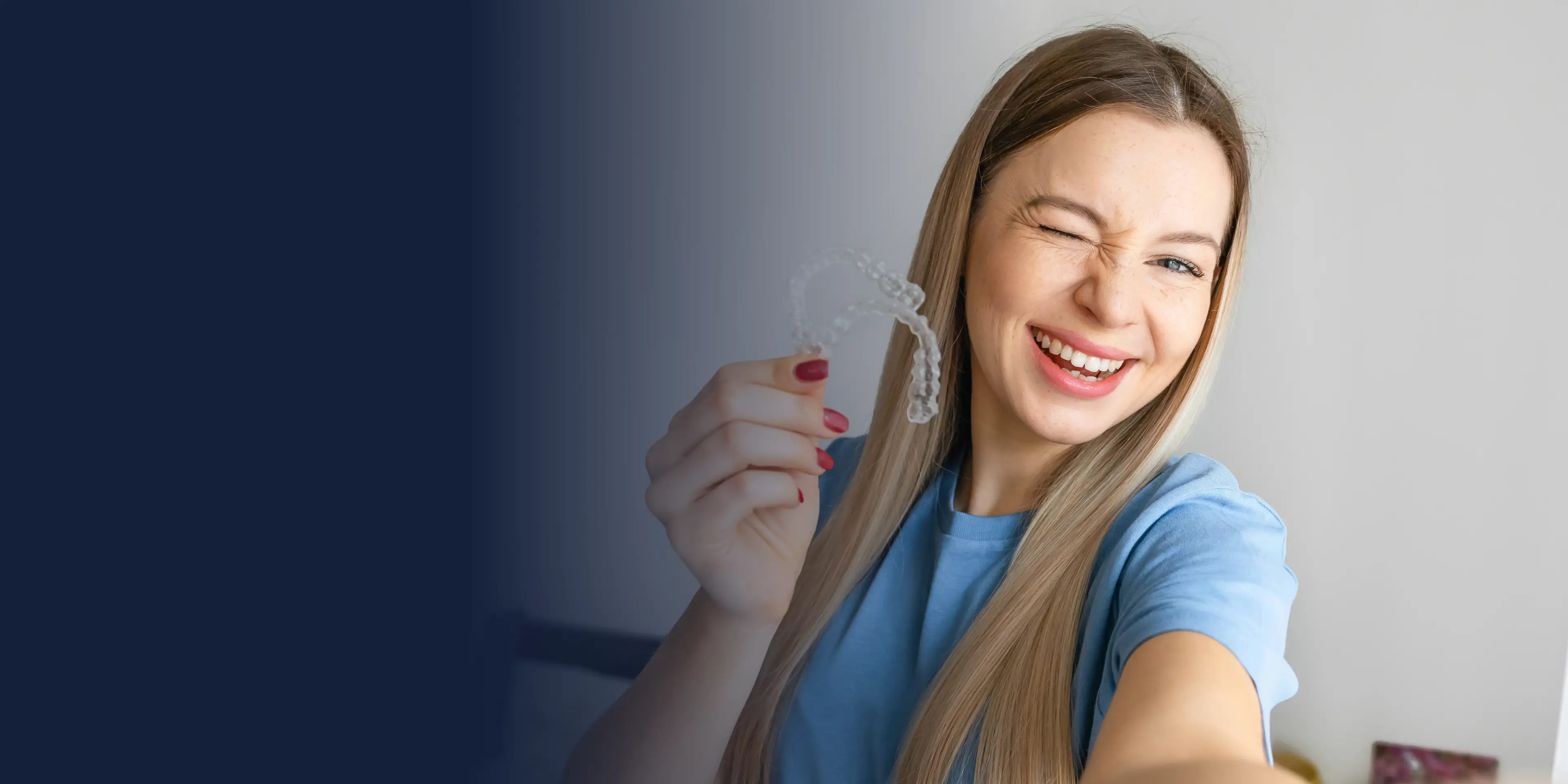 Woman smiling and winking while holding a clear dental aligner in her hand.