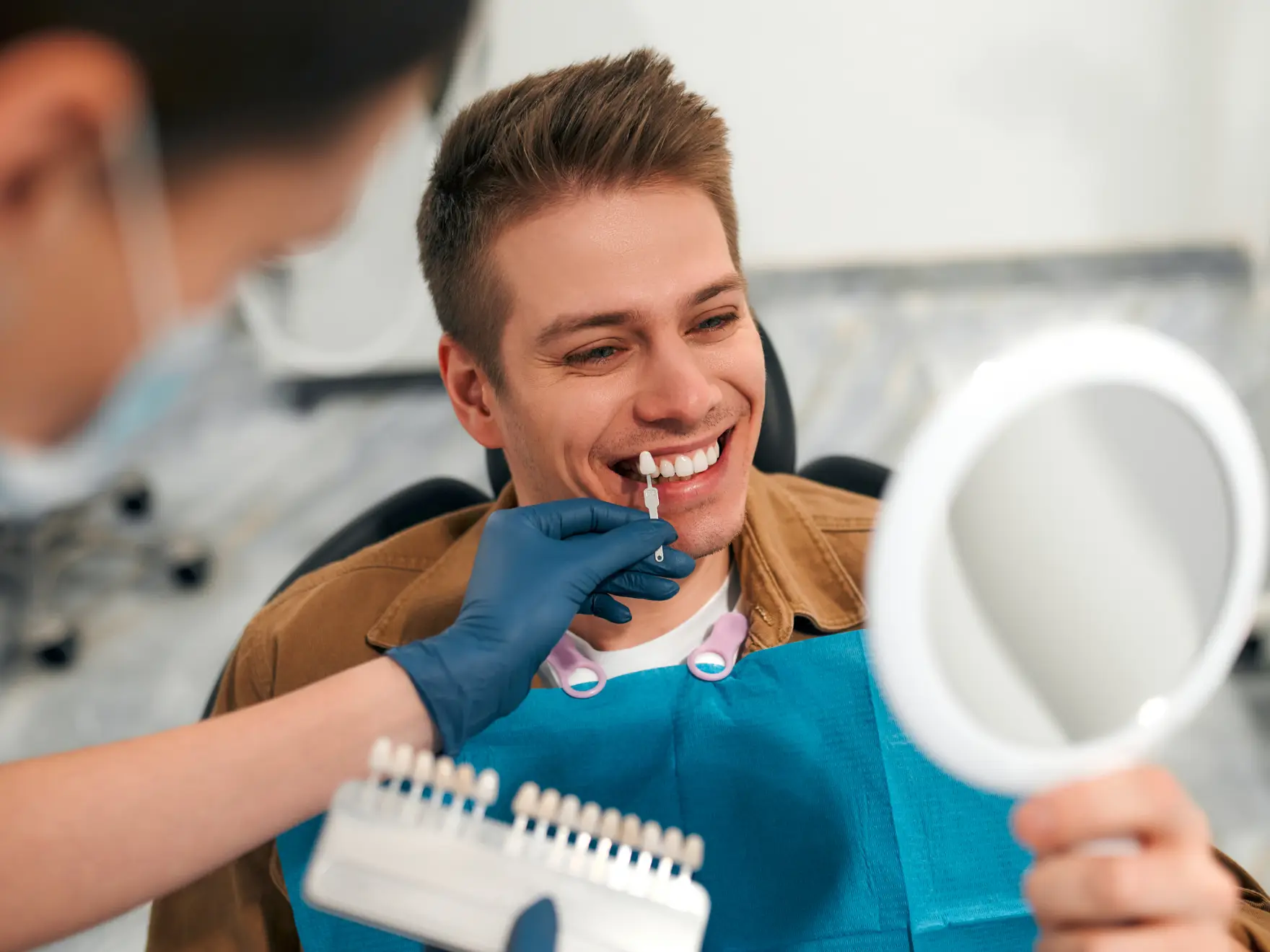 A person smiling holds a small mirror while a dental professional matches their teeth to a color chart.