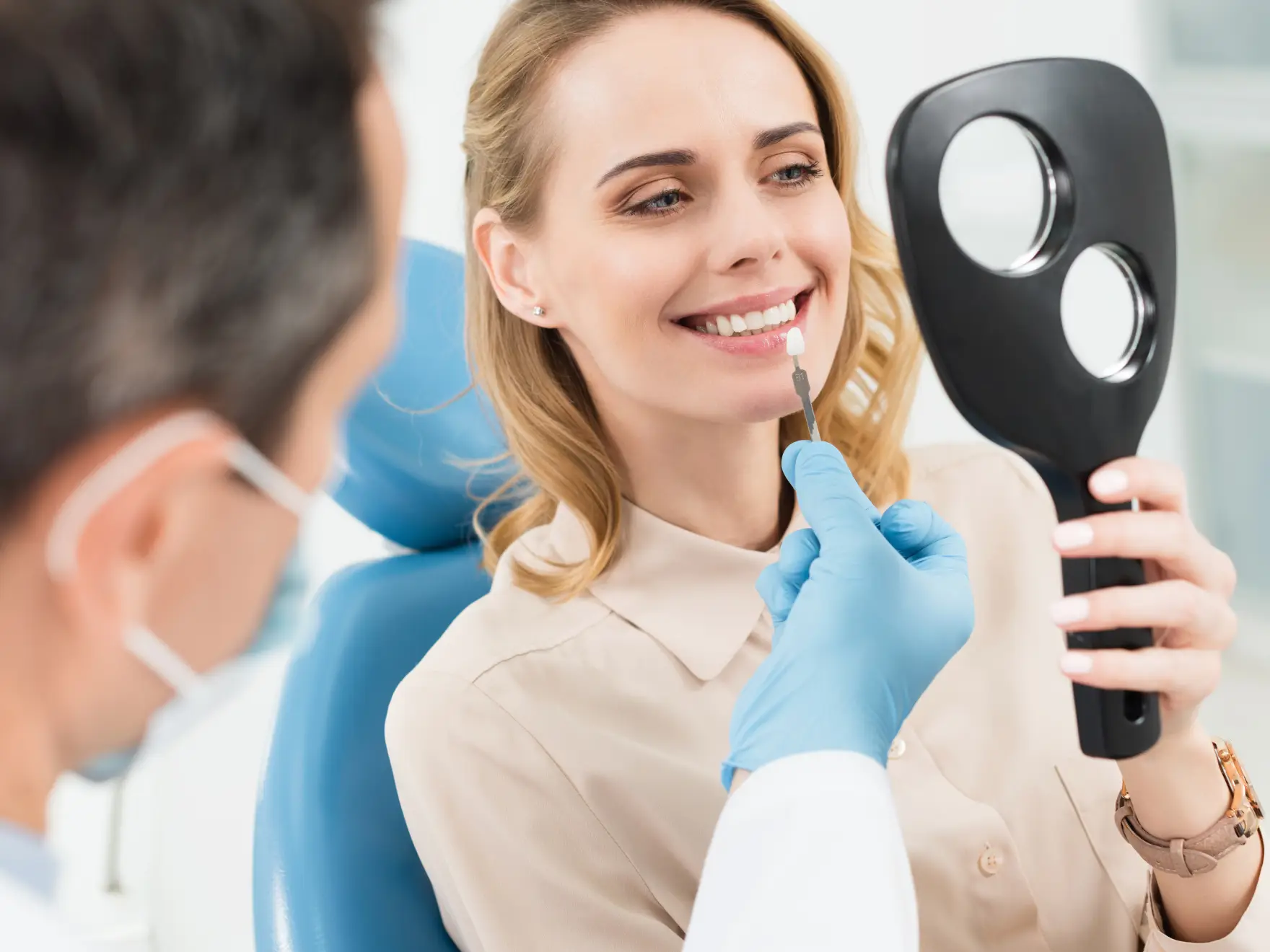 Dentist shows woman dental shade guide; she smiles while holding a mirror.