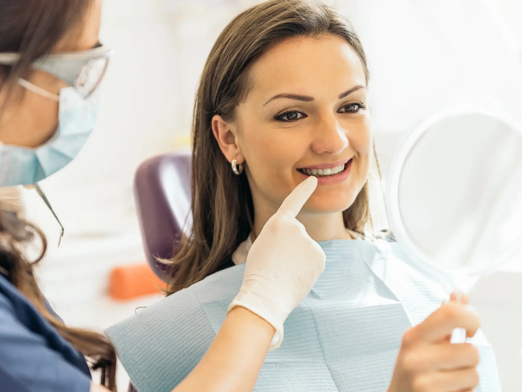 A dentist points at a woman's teeth as she examines them with a handheld mirror.