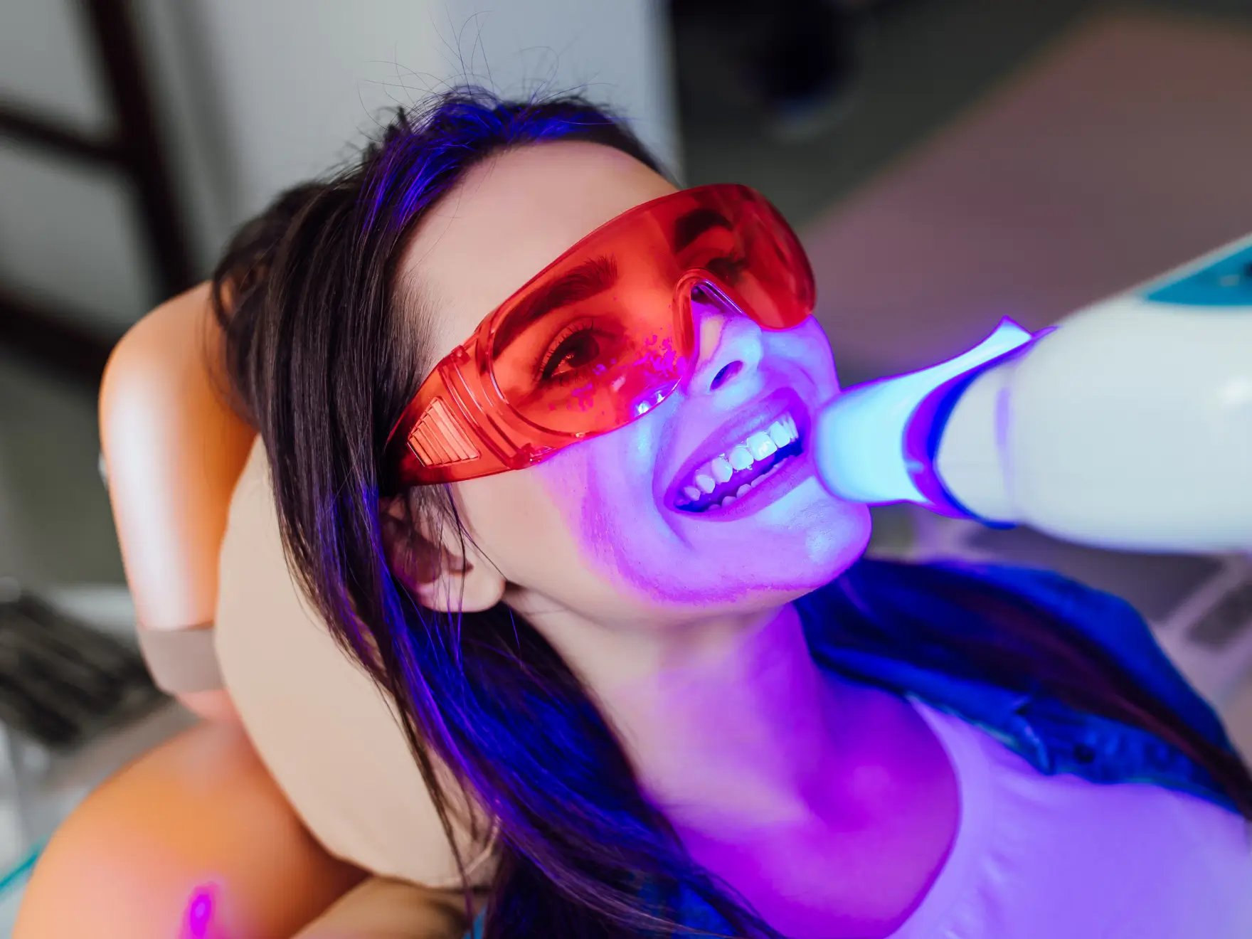 A woman sits in a dental chair undergoing a teeth whitening procedure with protective red glasses.